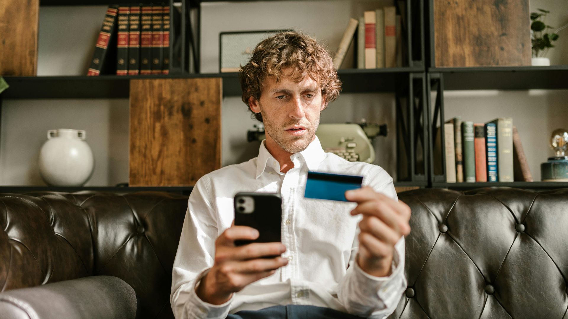 A man sitting on a leather sofa using a smartphone and credit card for online shopping.