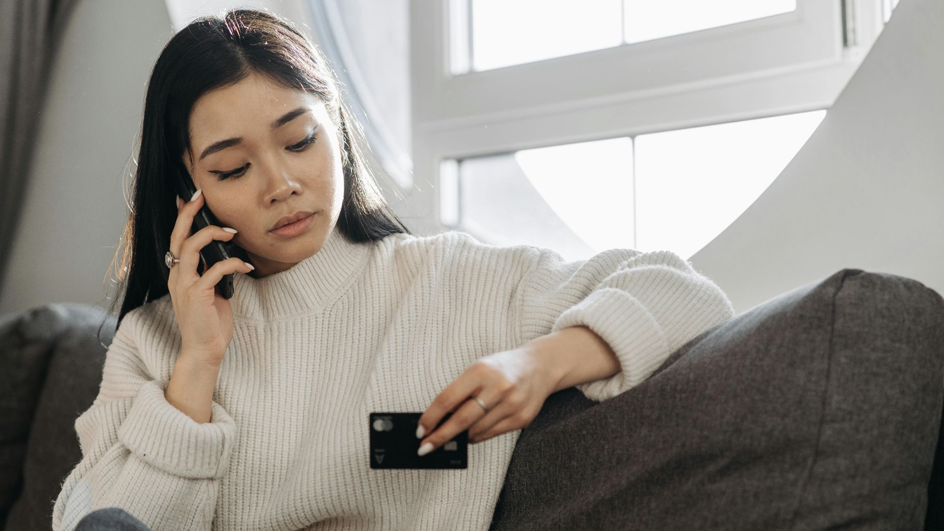 A woman sits on a sofa, multitasking with a phone and credit card, focusing on online shopping.