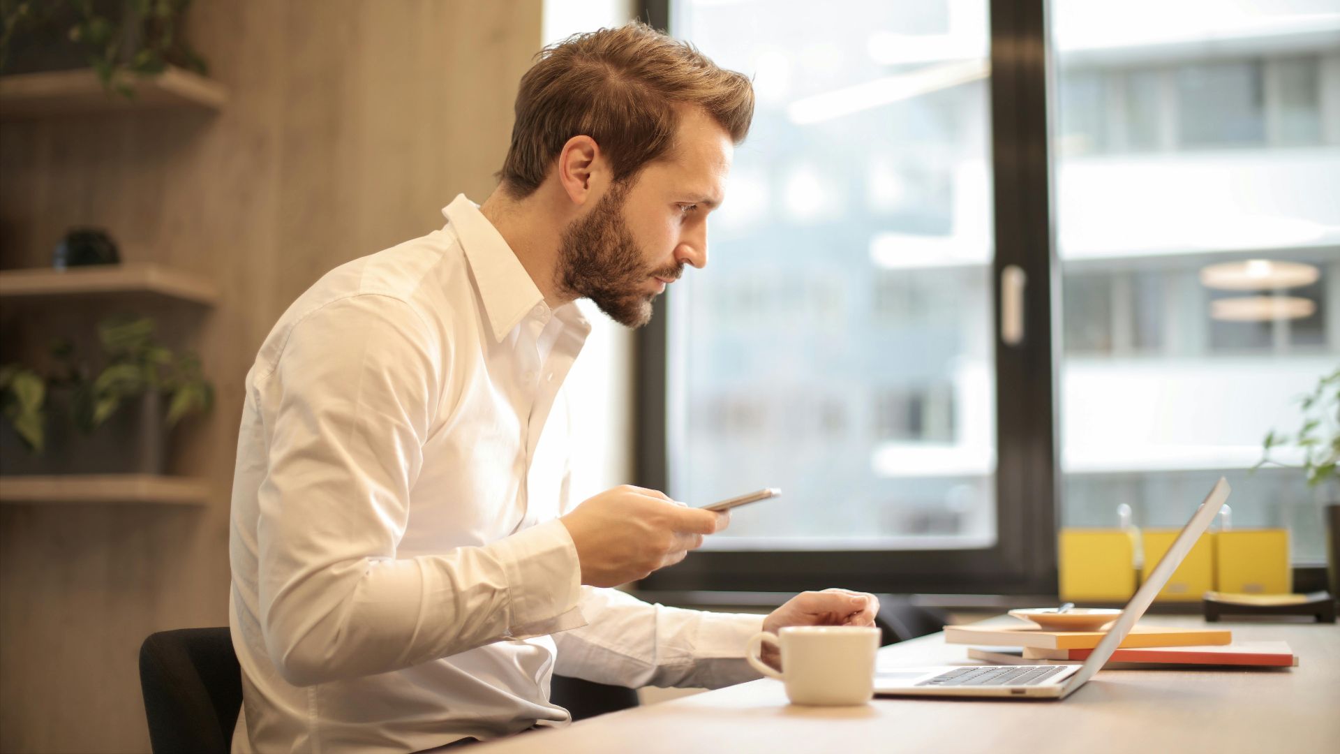 Focused businessman working on laptop while checking smartphone in modern office.