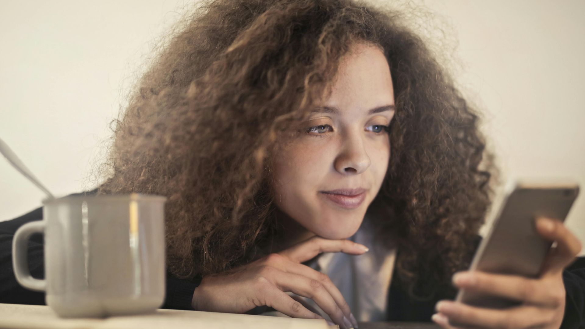 Young woman enjoying a relaxing moment while checking her smartphone indoors with a cup of coffee.