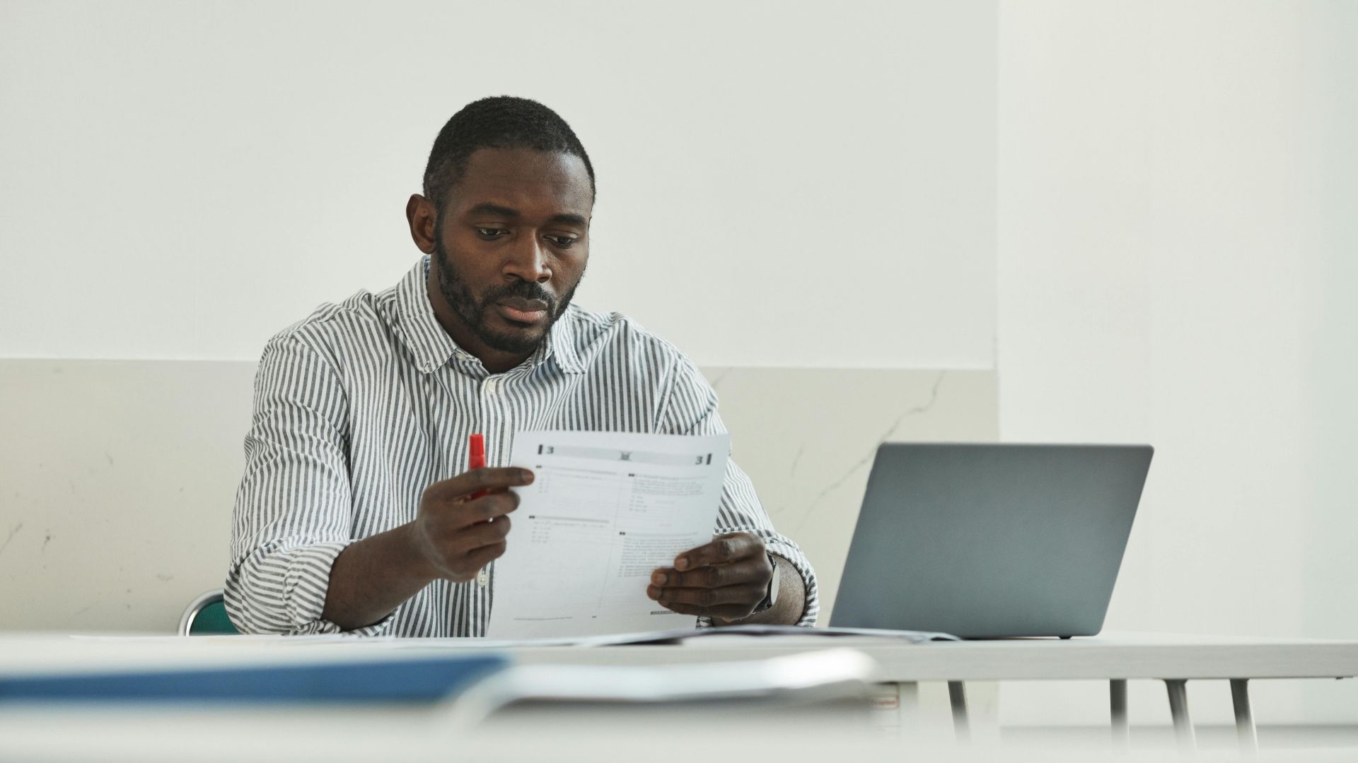 An adult man attentively grading test papers at his workspace with a laptop.