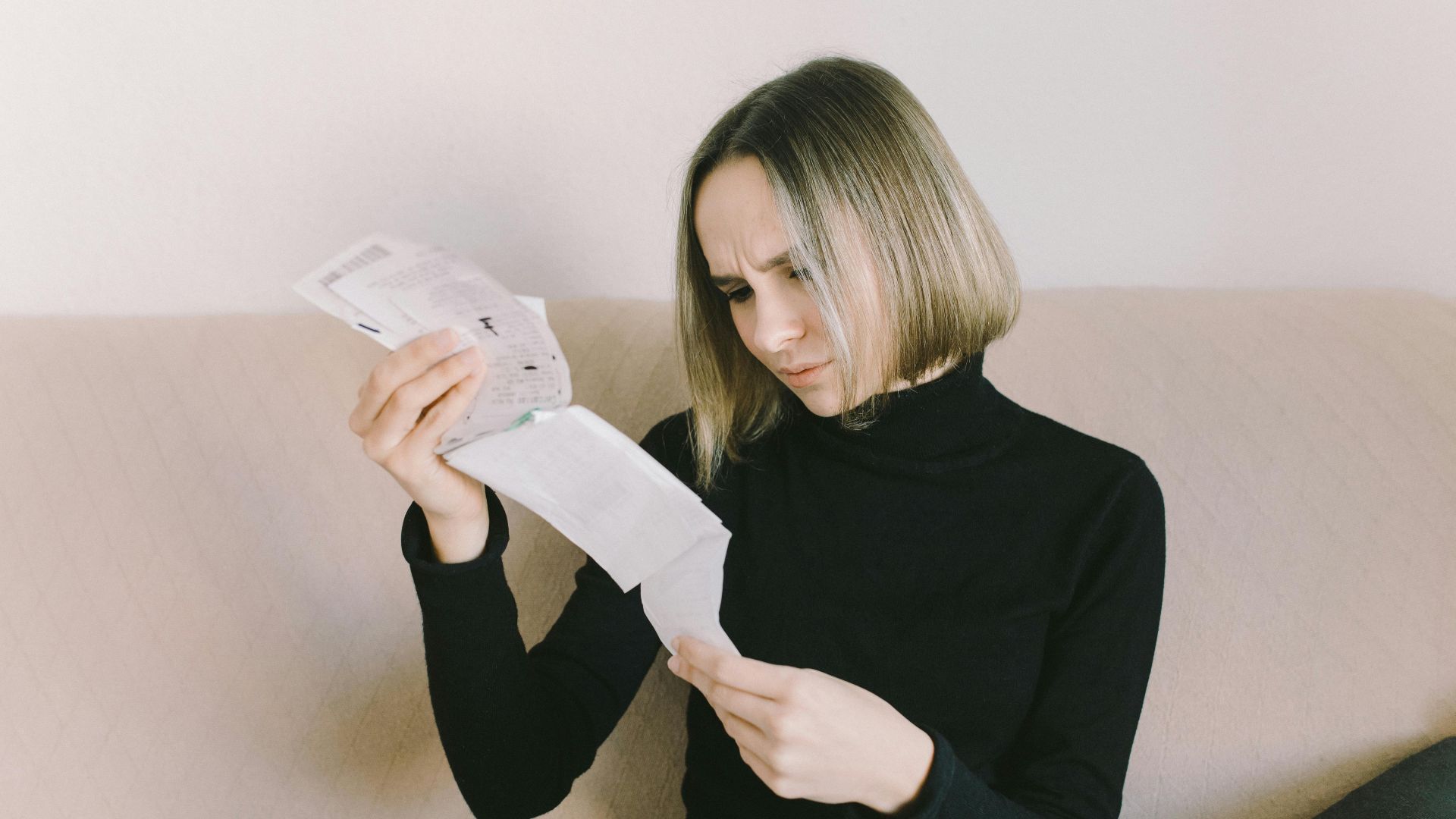 Concerned woman in black sweater examining bills on beige sofa indoors.