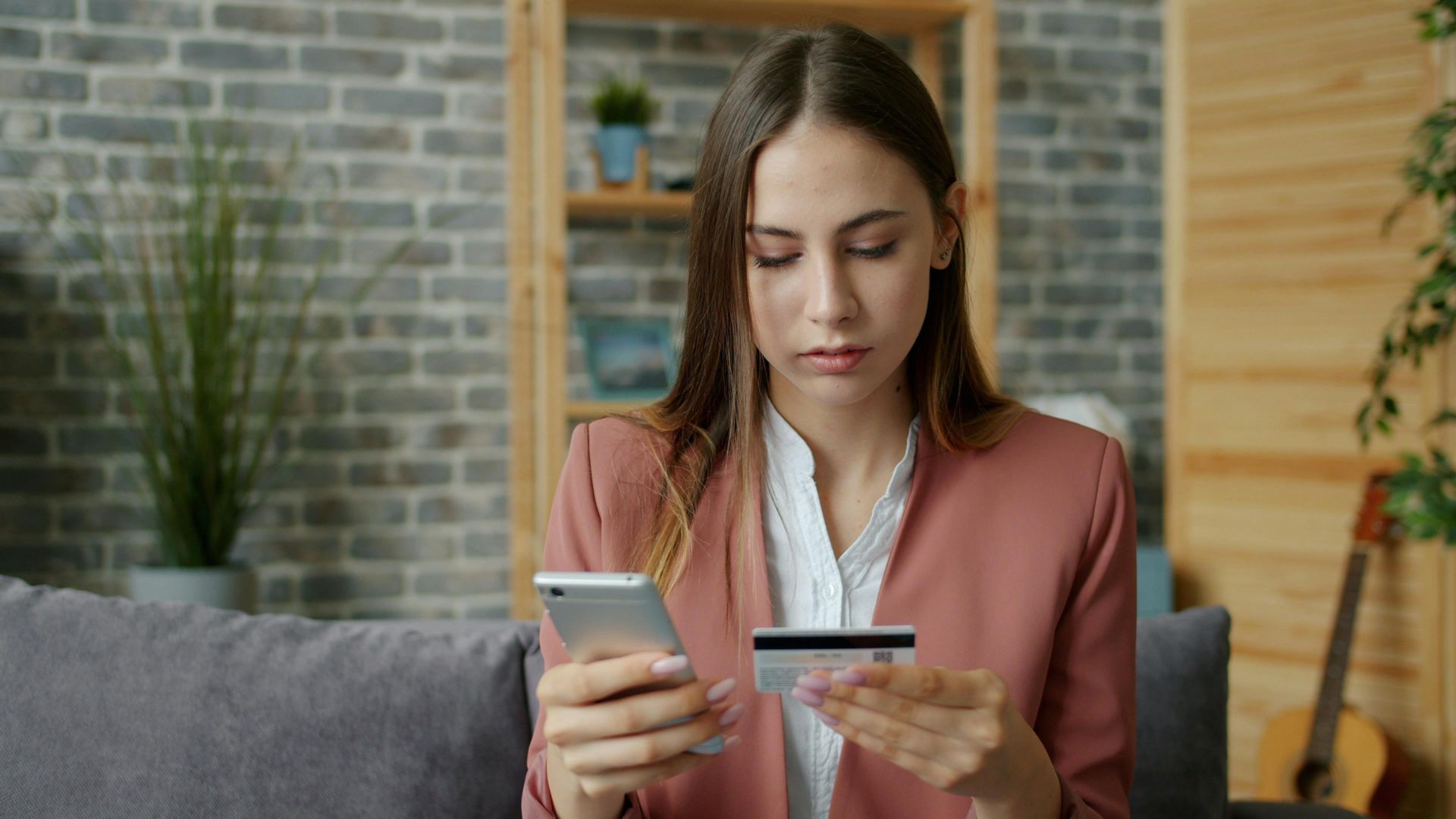 A young woman in a pink blazer shops online using a smartphone and credit card indoors.