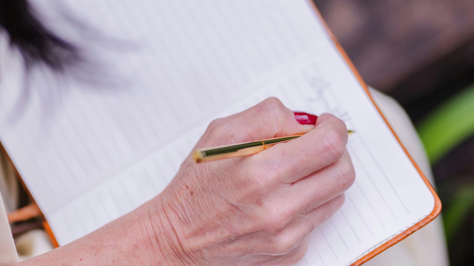 Close-up of a woman writing in a notebook while sitting outdoors, focusing on her hand and pen.
