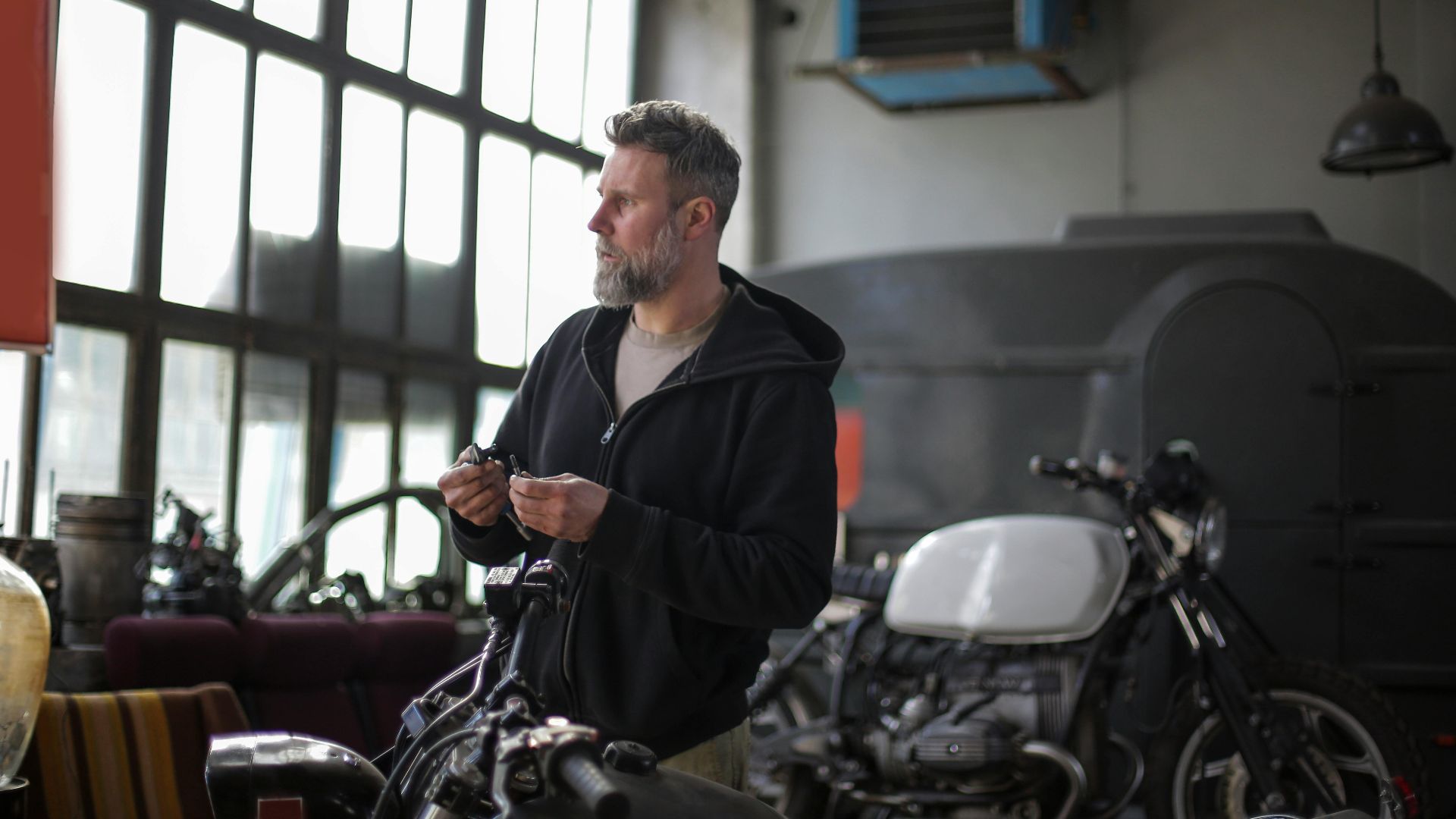 Side view of bearded man in casual wear standing in auto service holding detail in hand and looking away while repairing motorbike