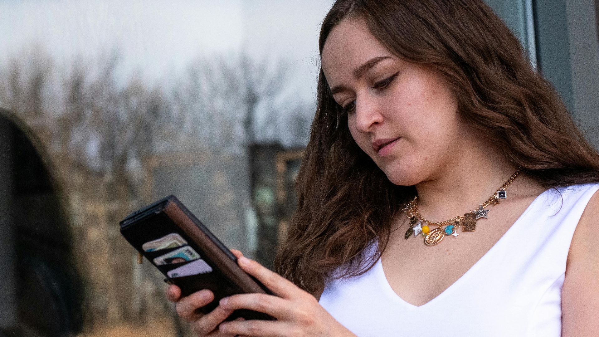 A woman in a white dress stands by a reflective window using her smartphone outdoors.