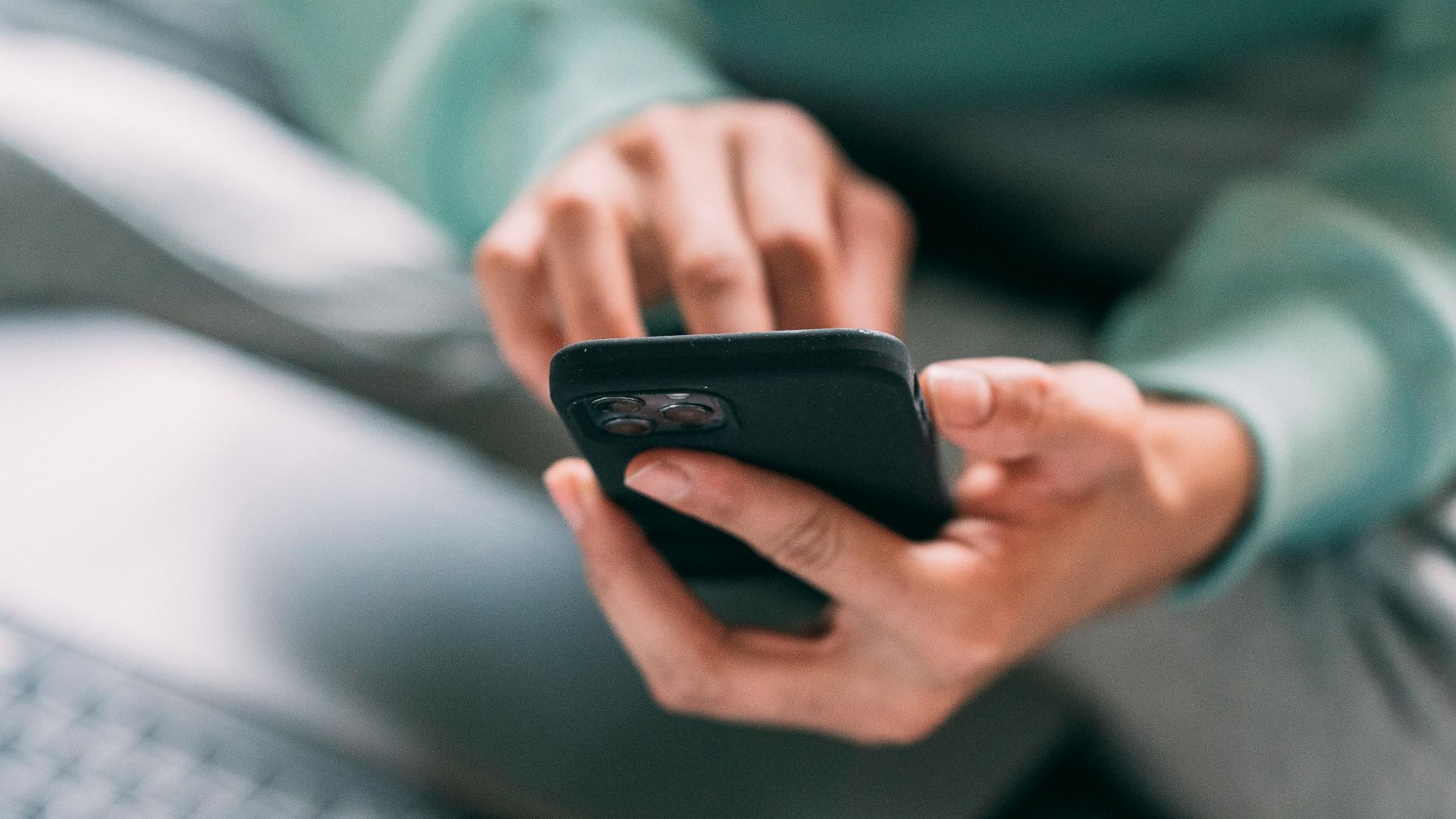 Crop anonymous male freelancer text messaging on modern cellphone while sitting on bed with laptop during remote work at home