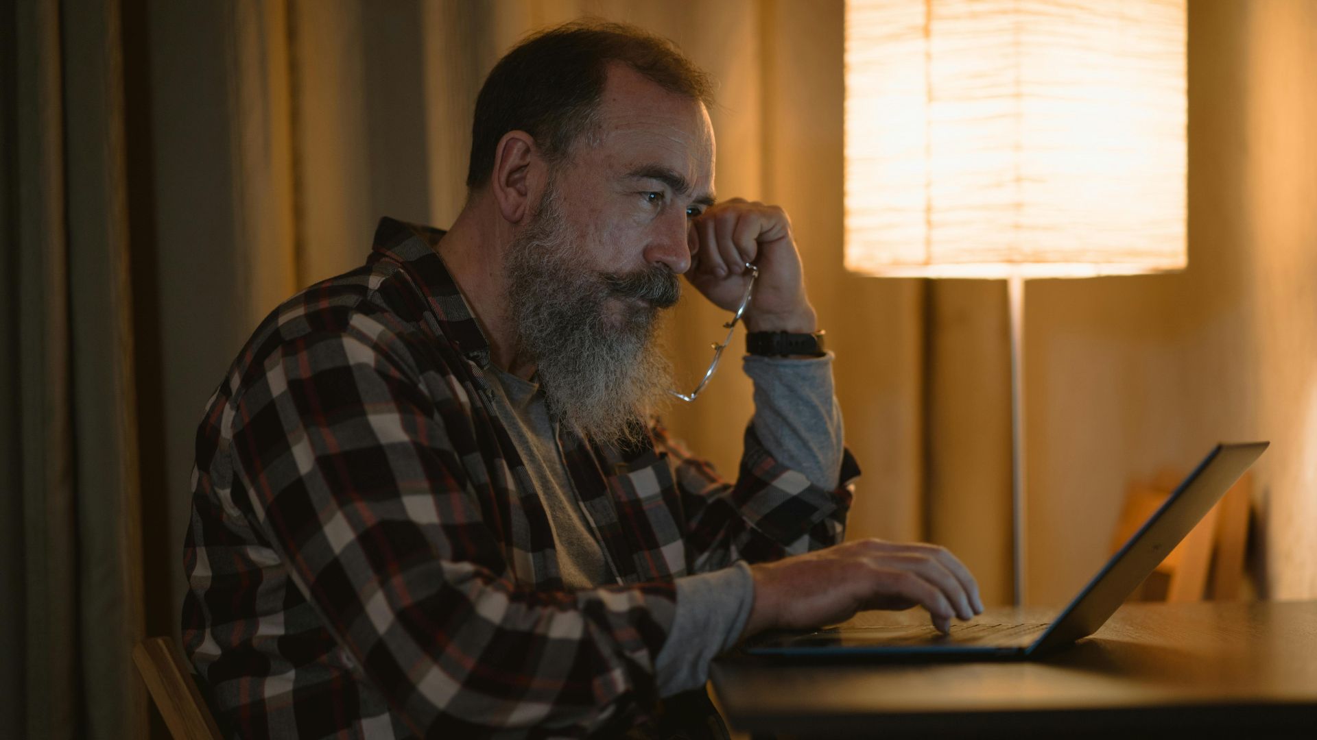Elderly man with a beard working late on laptop under warm lamp light indoors.
