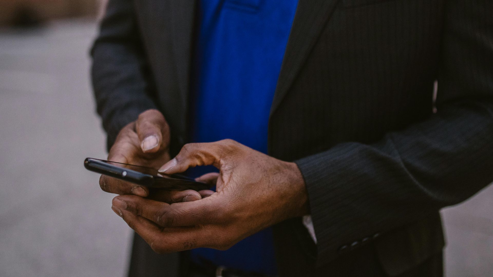 A man in a black suit using a smartphone while standing outdoors during the day.