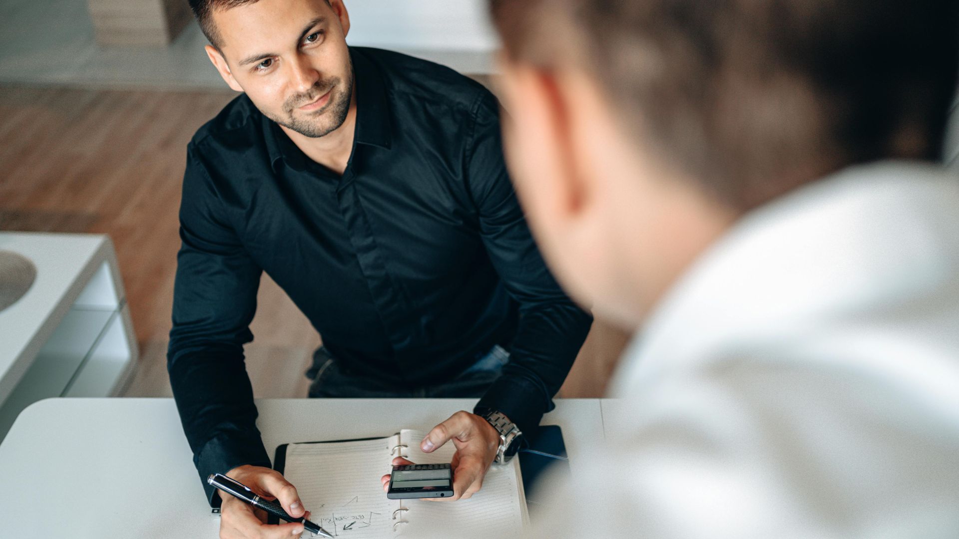 Two men in a business meeting indoors, discussing and taking notes.