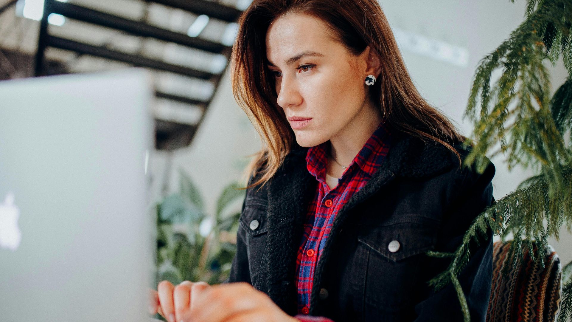 Young woman in plaid shirt intensely working on laptop in a modern environment with greenery.