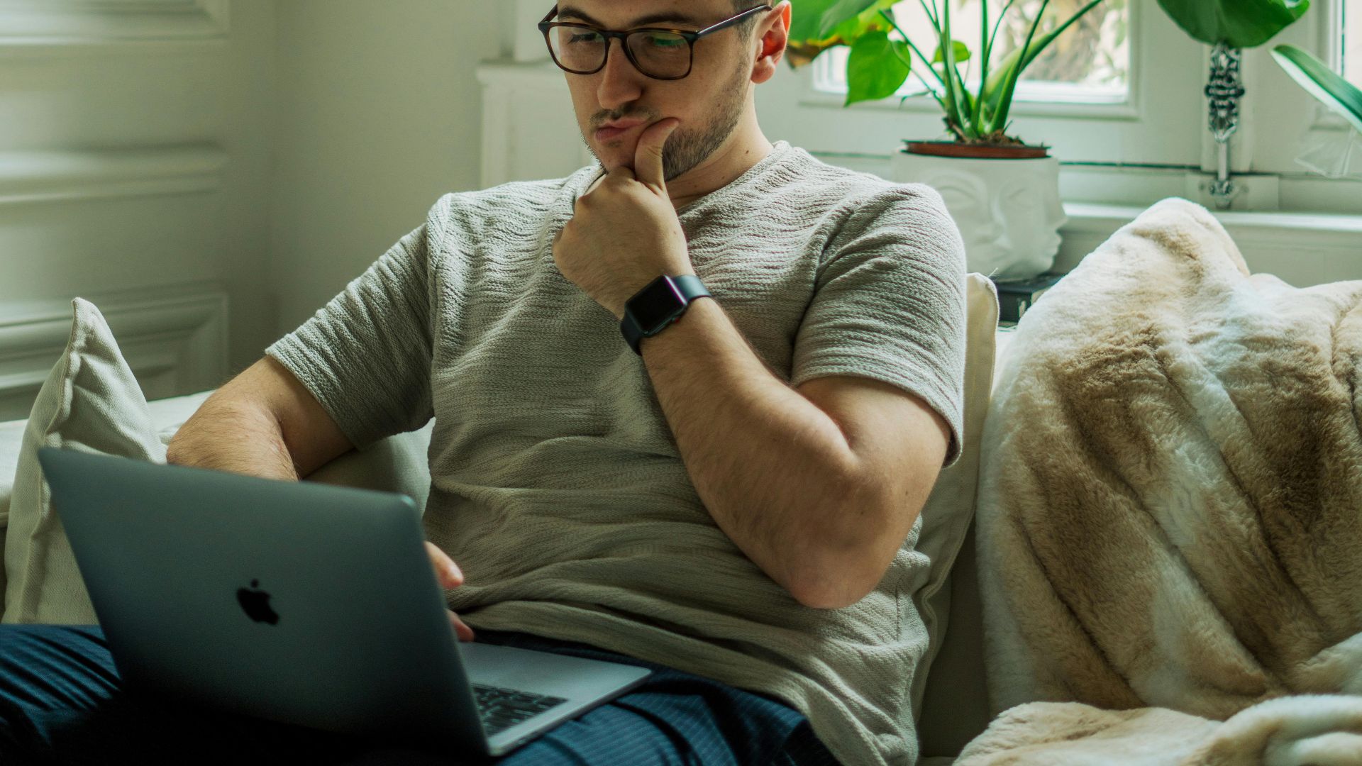 A man wearing eyeglasses using a laptop in a cozy home setting with natural light.
