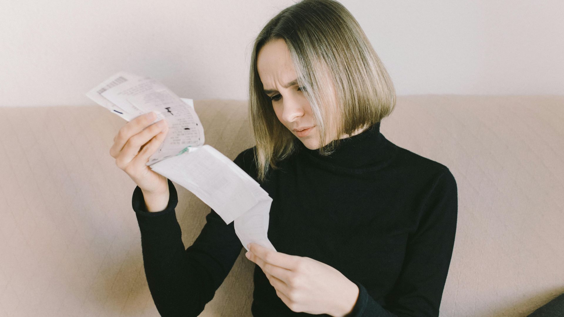 Concerned woman in black sweater examining bills on beige sofa indoors.
