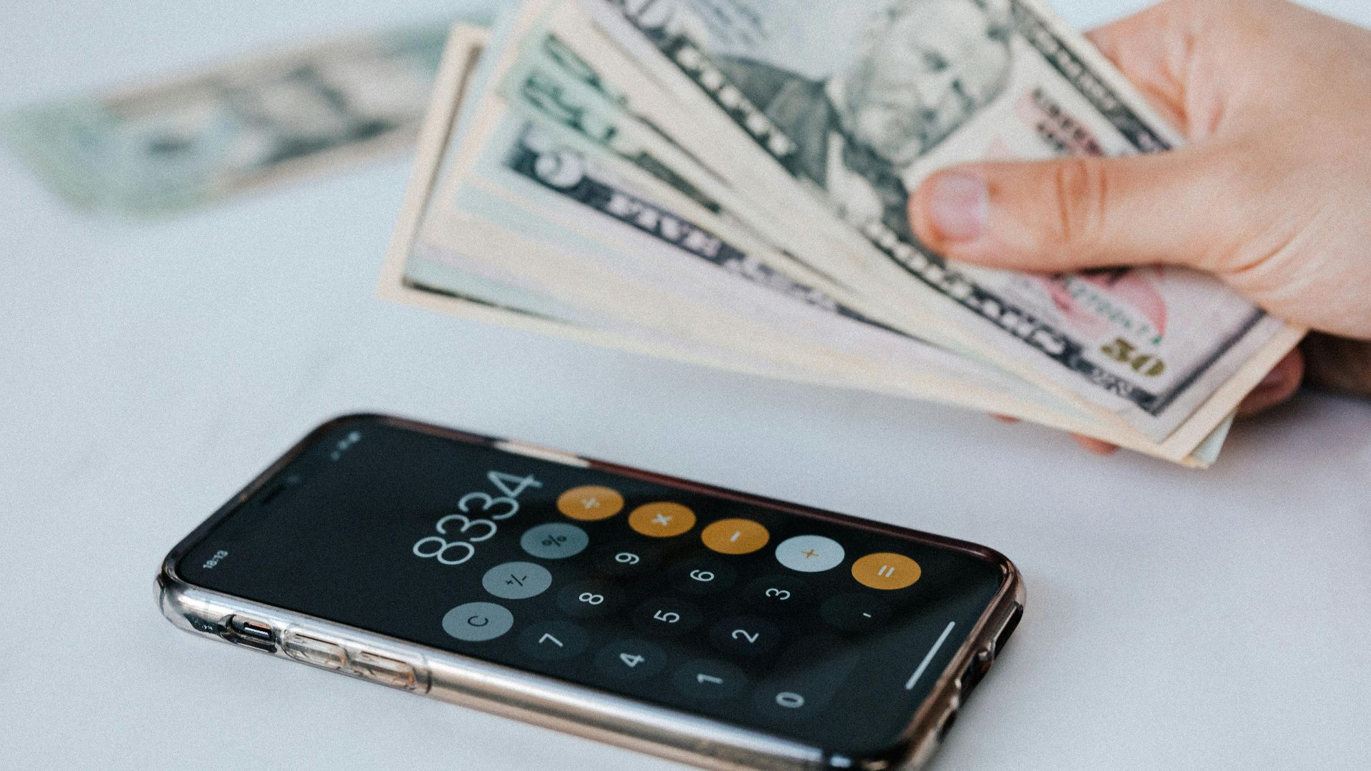 Calculator application on mobile phone screen on white surface near crop anonymous person holding pile of dollars