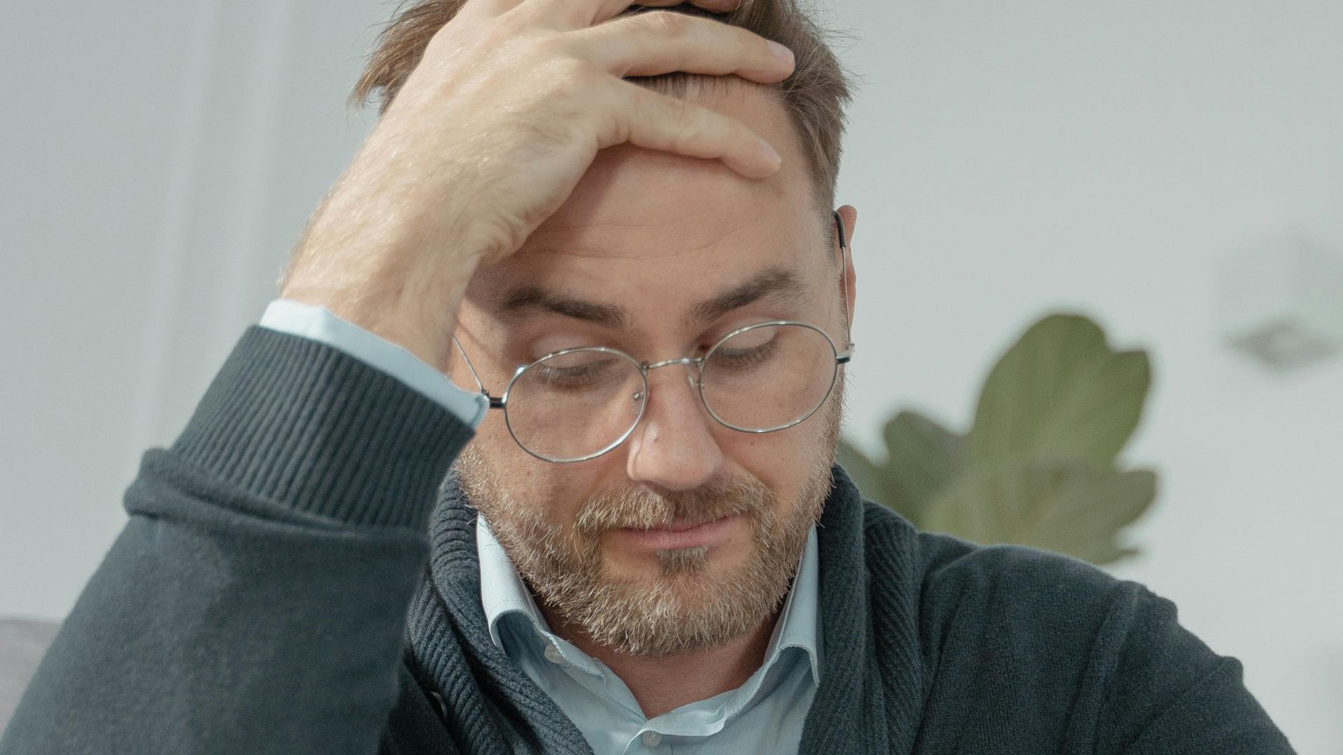 A thoughtful man with glasses sits indoors, hand on head, reflecting stress or contemplation.