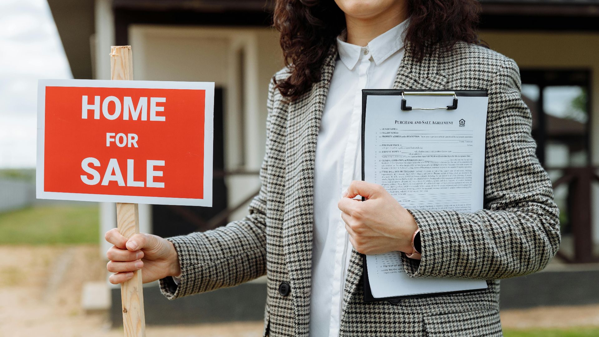 A real estate agent holding a home for sale sign and clipboard outside a property.
