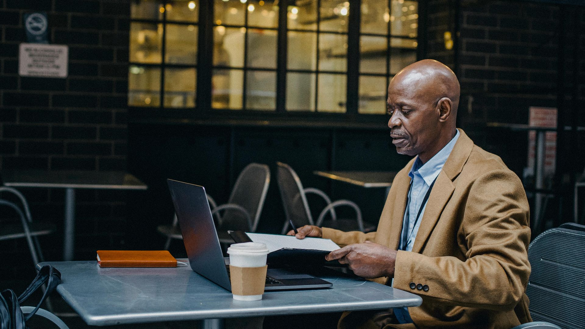 A middle-aged man in a café working on a laptop with a coffee and notebook.