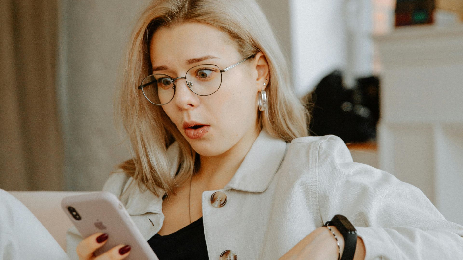 Surprised woman with glasses reacts to news on her phone while relaxing indoors.