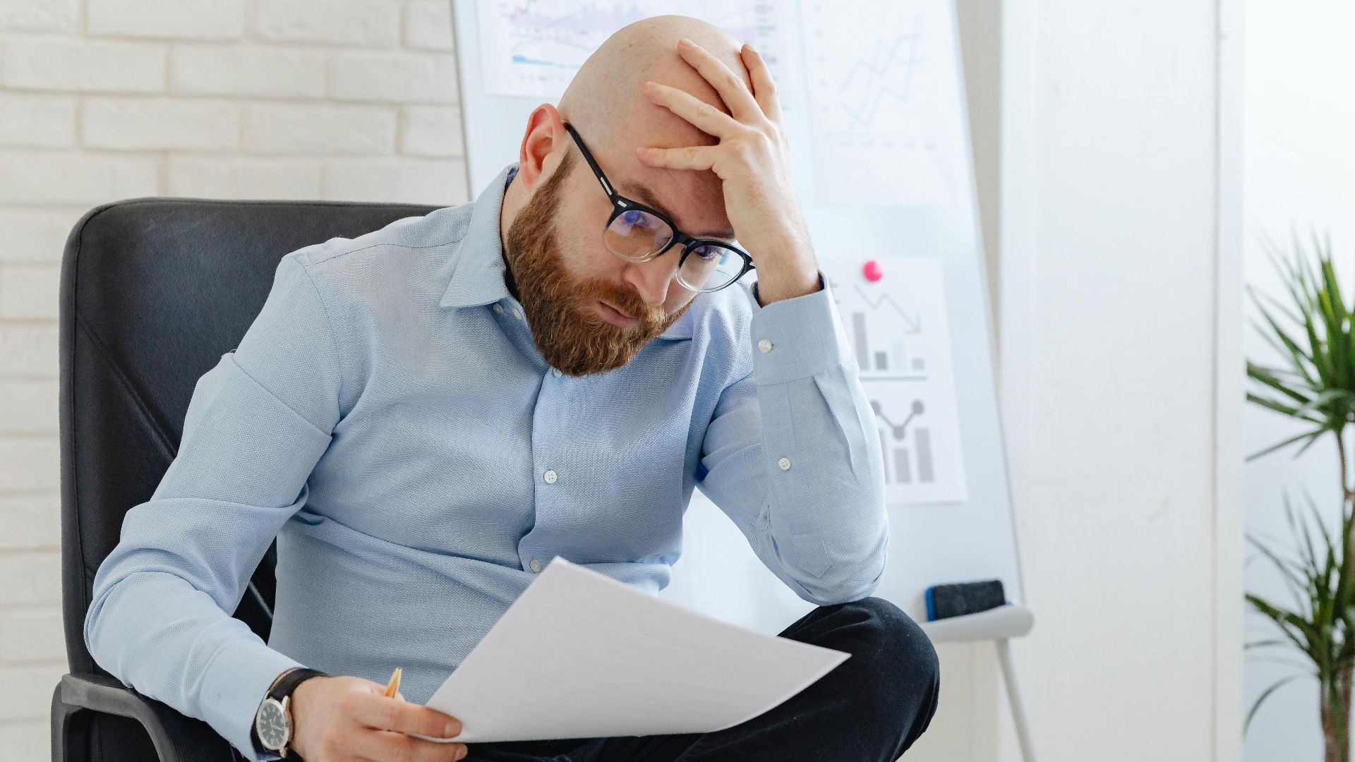 Bald man with glasses sitting in office looking worried at document.