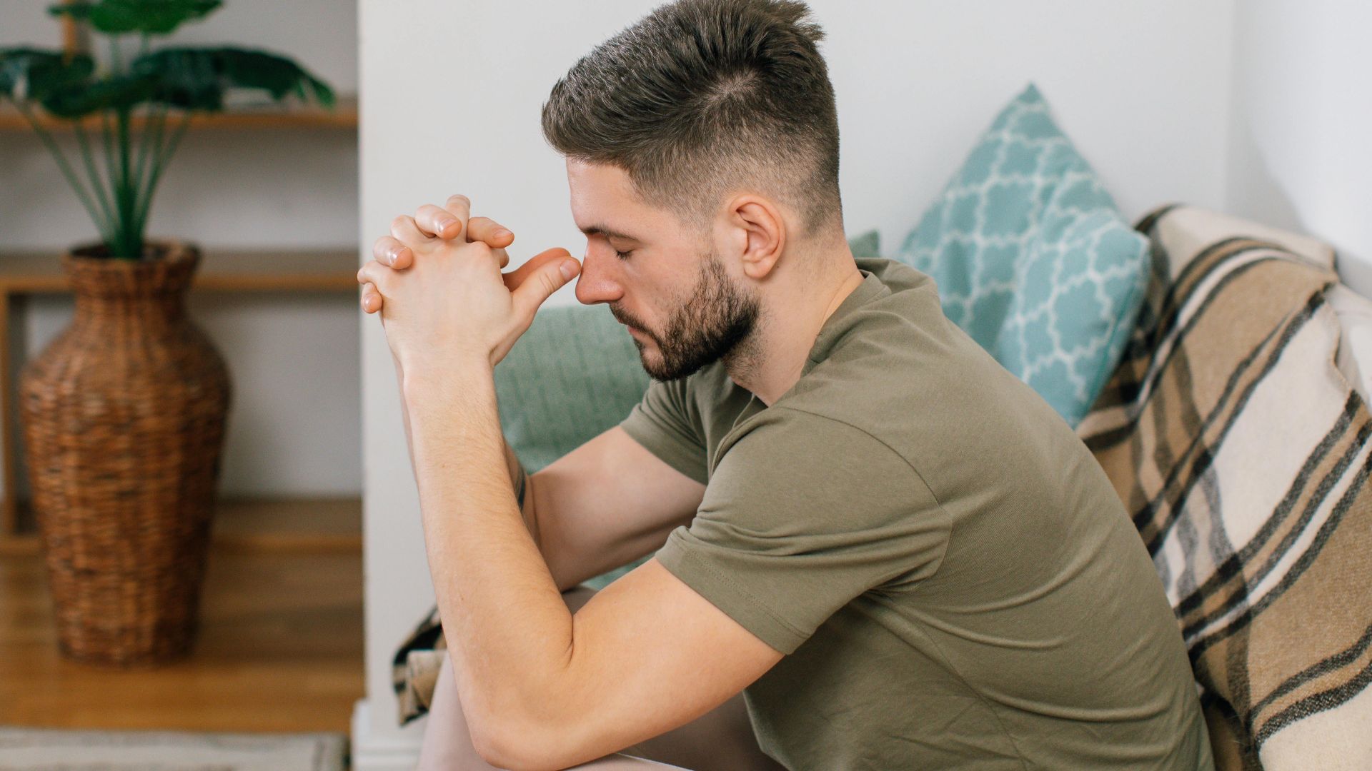 A young man with clasped hands, sitting thoughtfully on a sofa in a cozy living room.