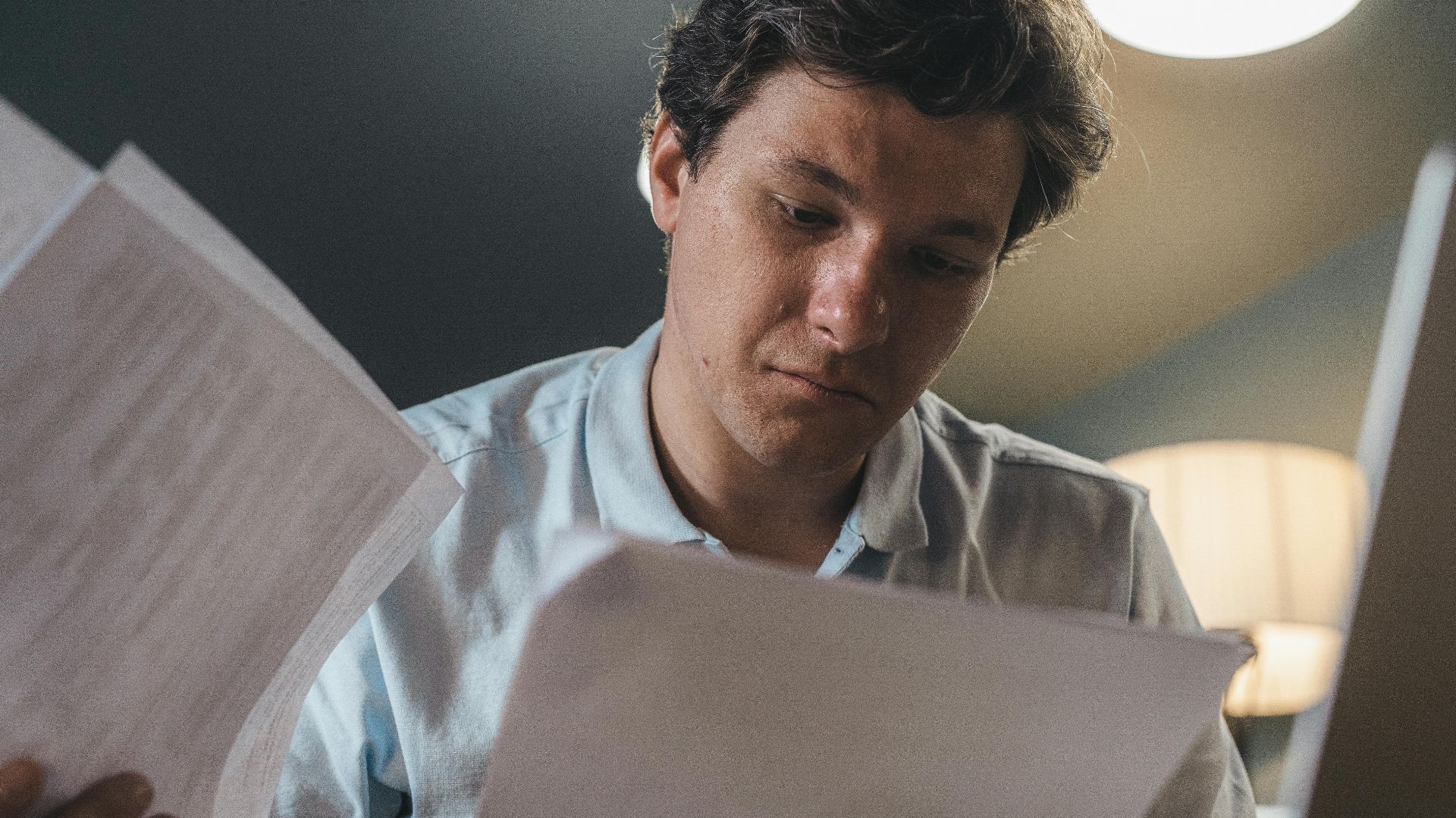 Serious man wearing a light blue shirt intently reading printed papers indoors under warm lighting.