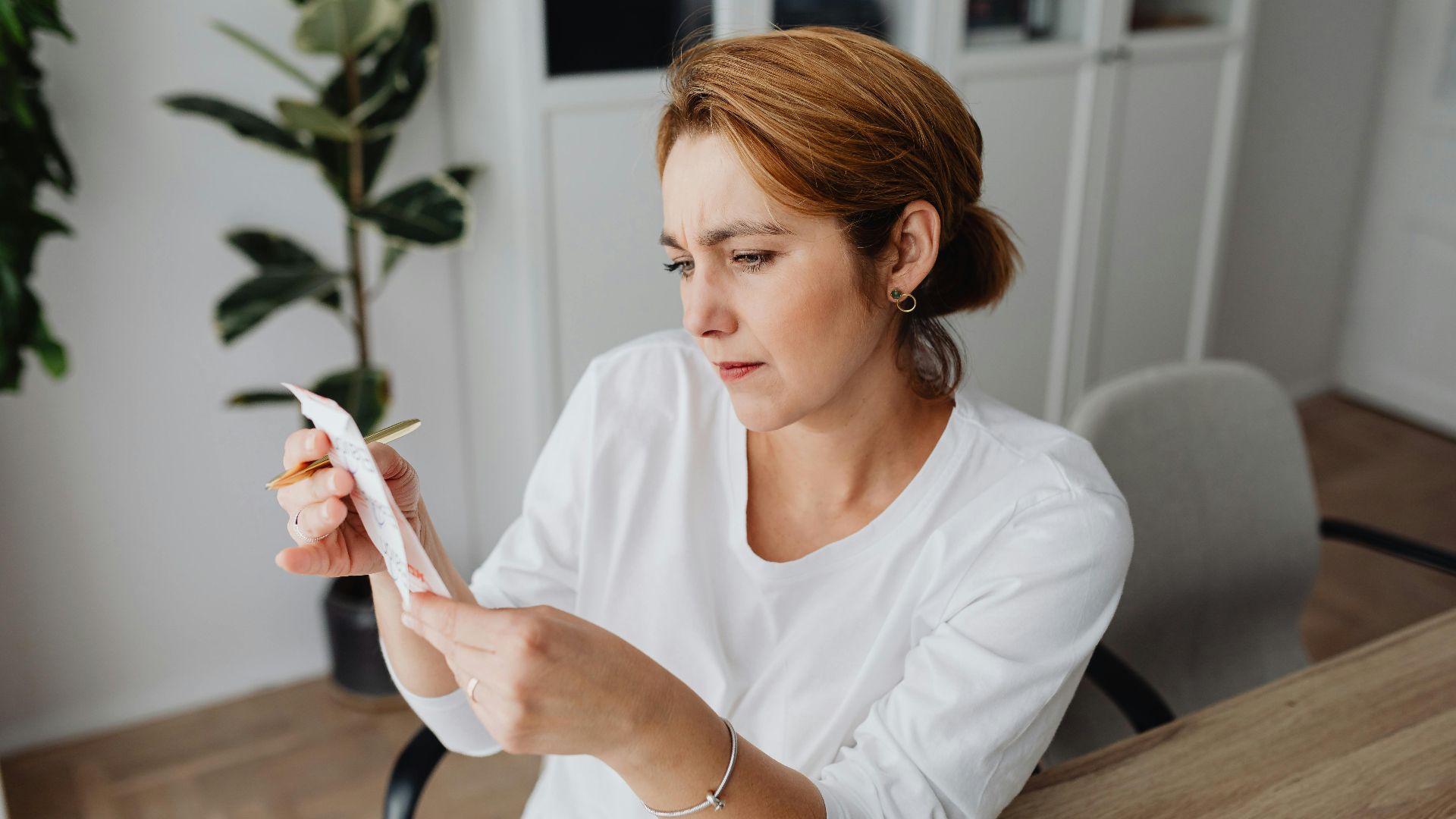 A concerned adult woman examines a receipt while sitting at her office desk, indicating financial scrutiny.