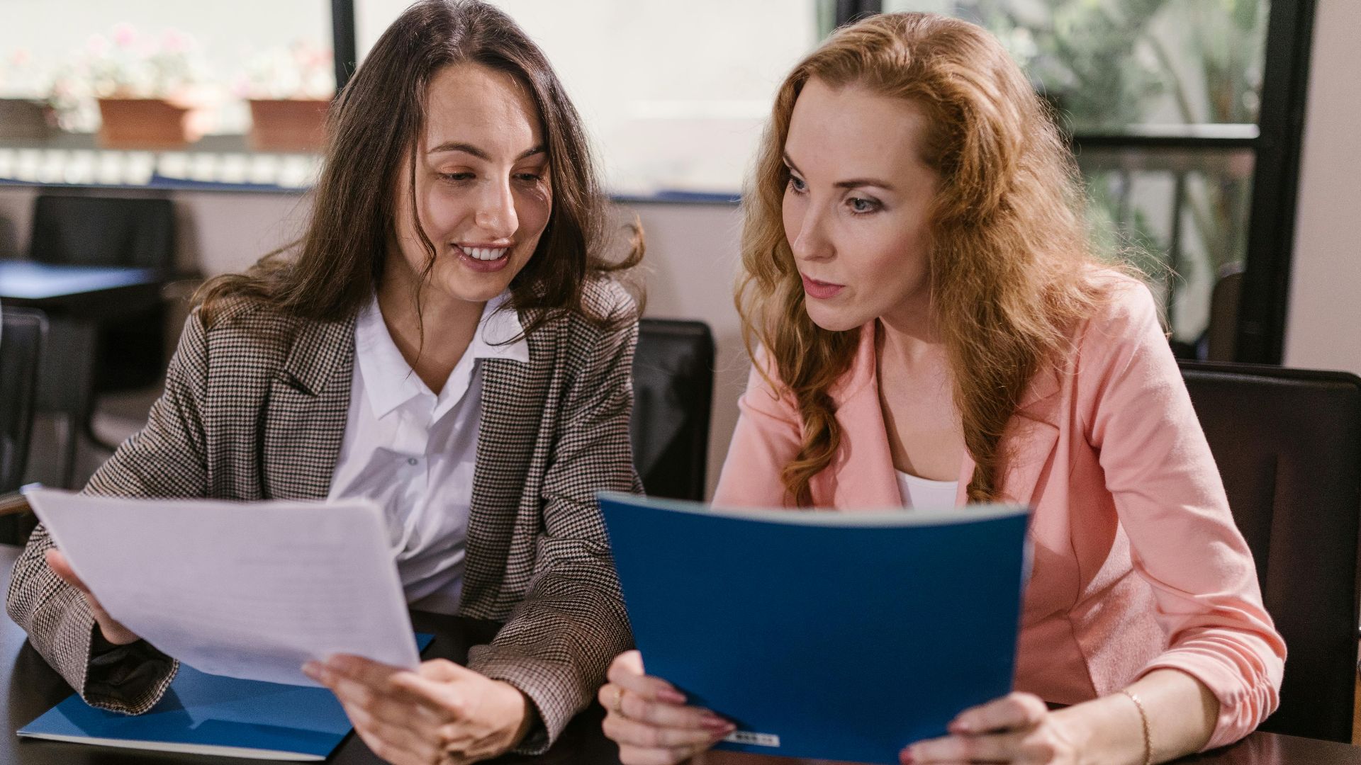 Two businesswomen reviewing documents together in an office setting, emphasizing teamwork and cooperation.