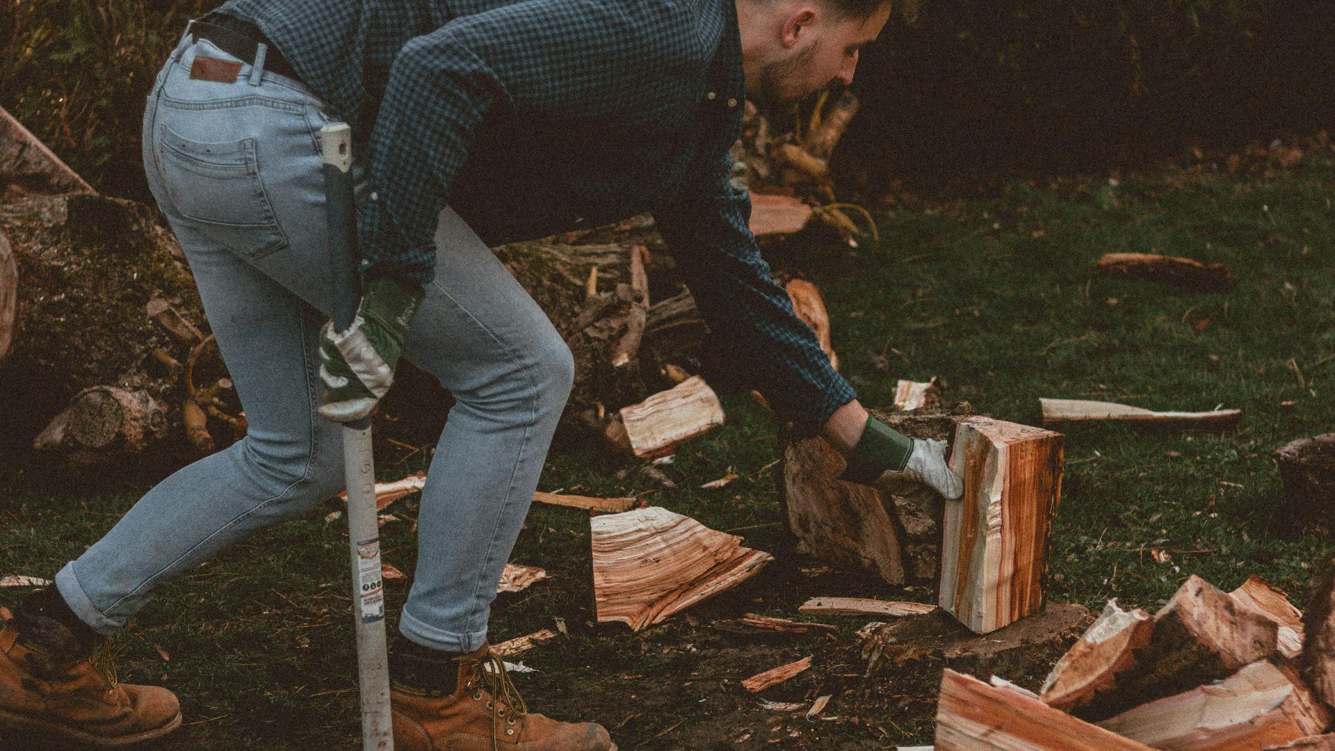 A man chops firewood with an axe in a grassy outdoor setting, surrounded by split logs during a summer evening.