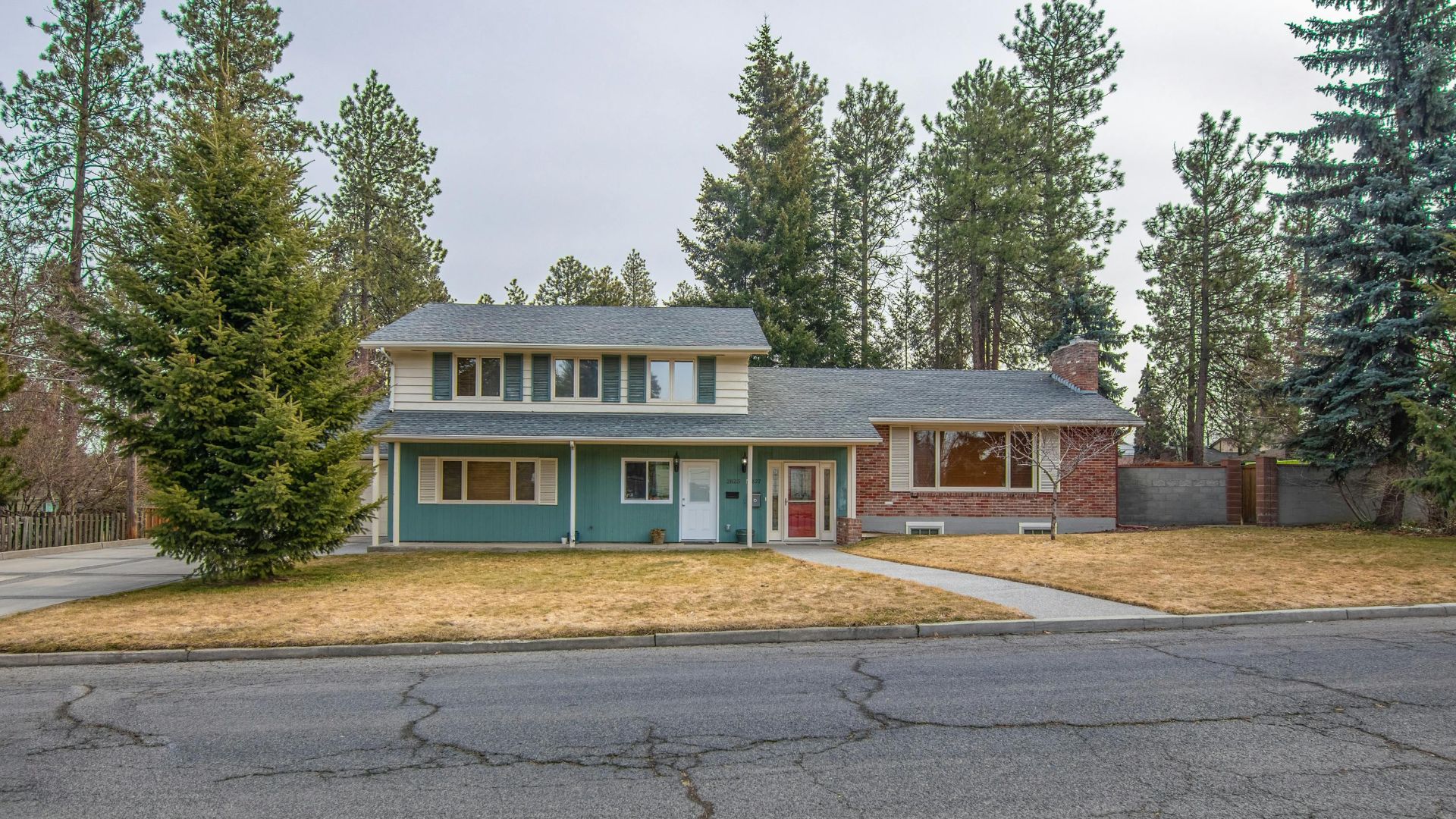 Two-story suburban house with green siding and brick accents, surrounded by trees.
