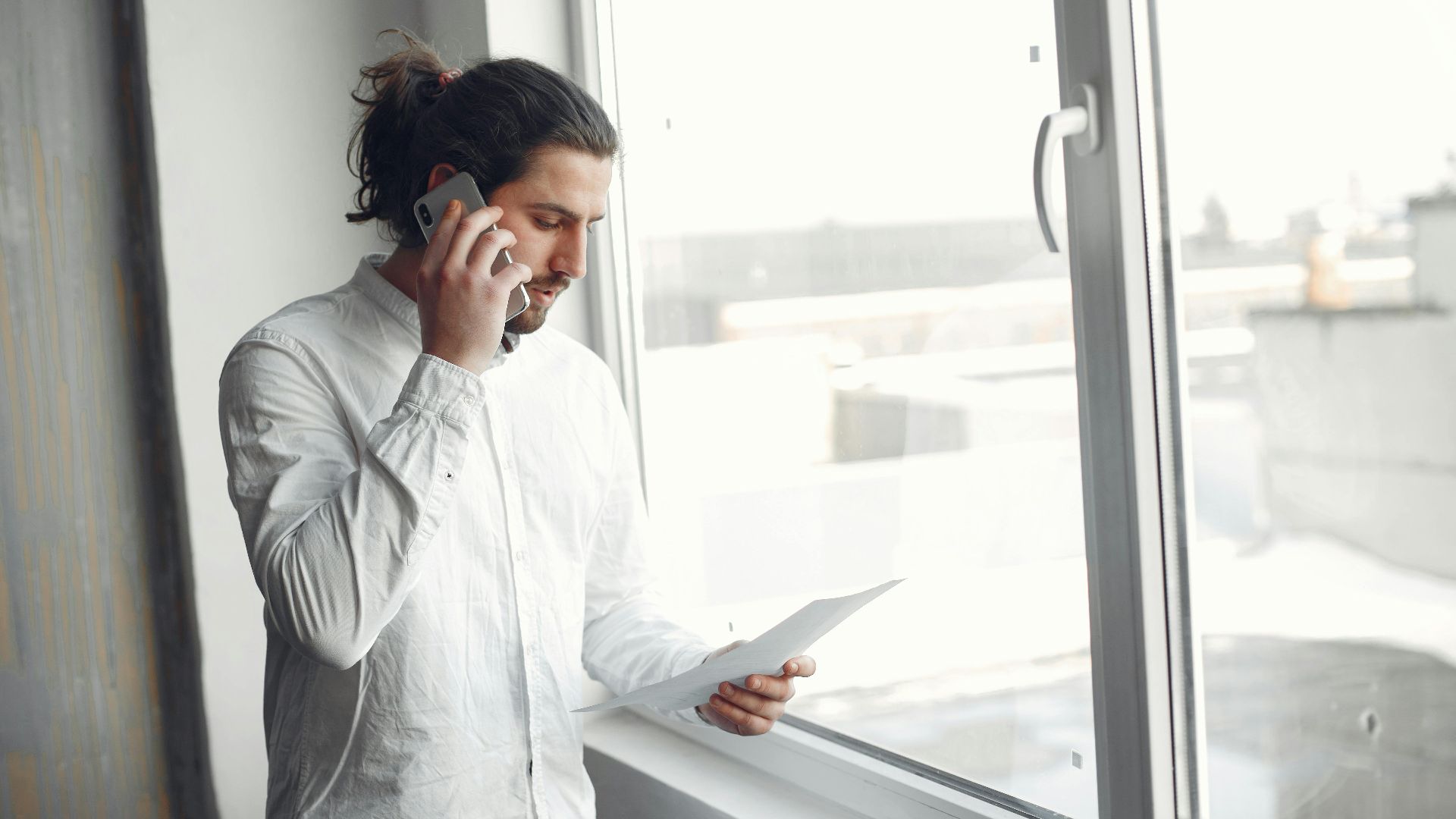 Young man in white shirt, on phone call holding a document, standing by a large window.