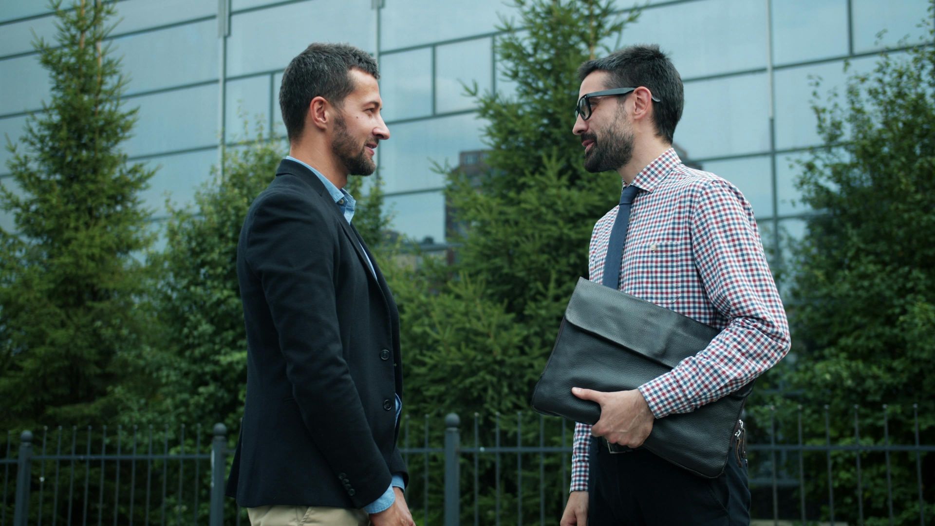 Two businessmen engage in conversation outdoors, standing beside a modern building.