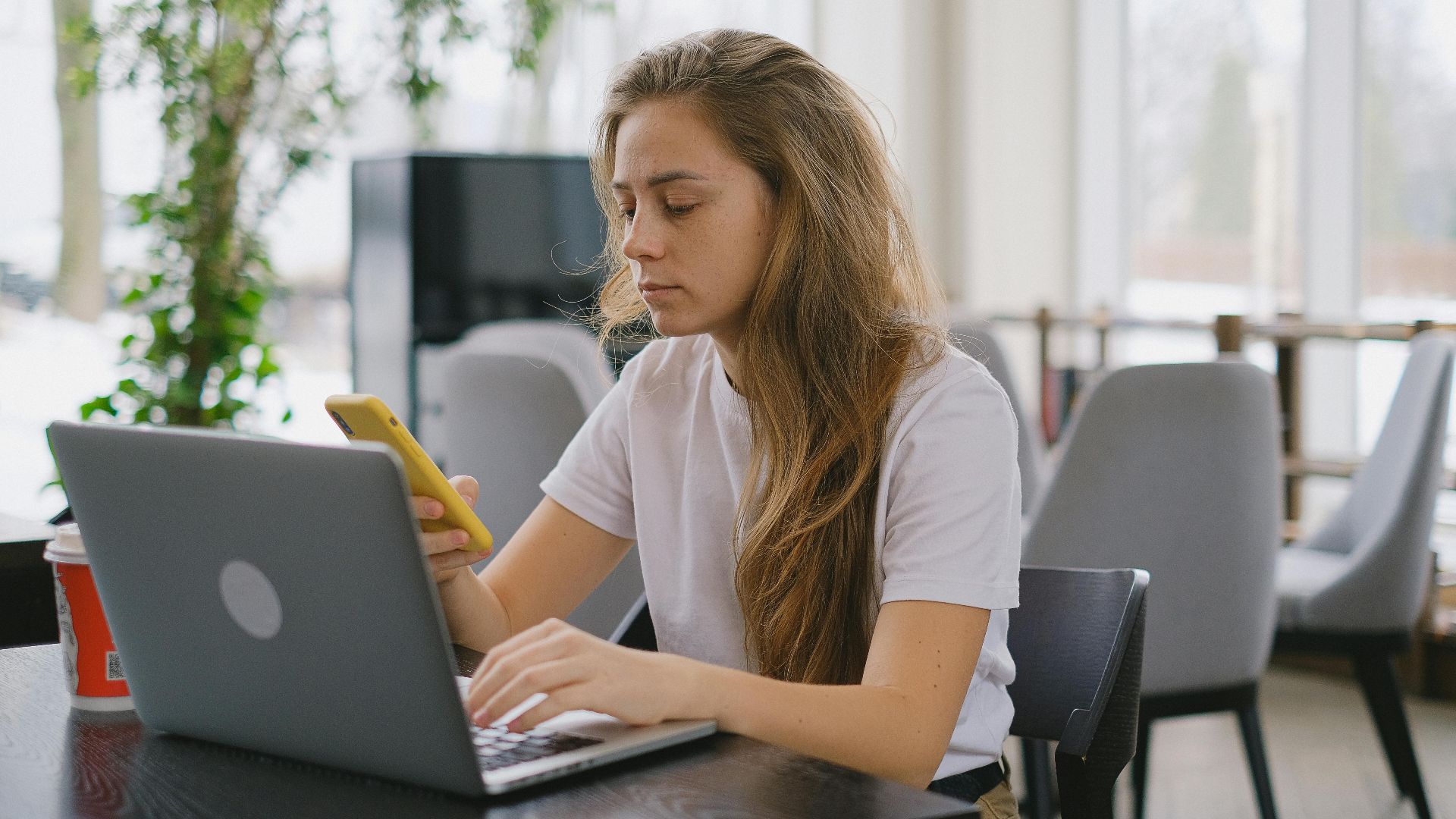 Focused woman using a laptop and smartphone at a cafe, showcasing modern multitasking.