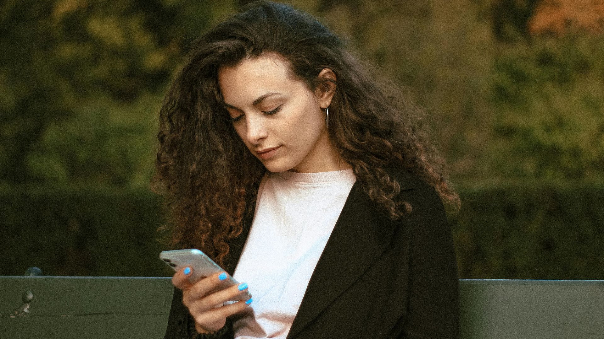 Woman with curly hair checking smartphone while sitting outdoors in Warsaw park.