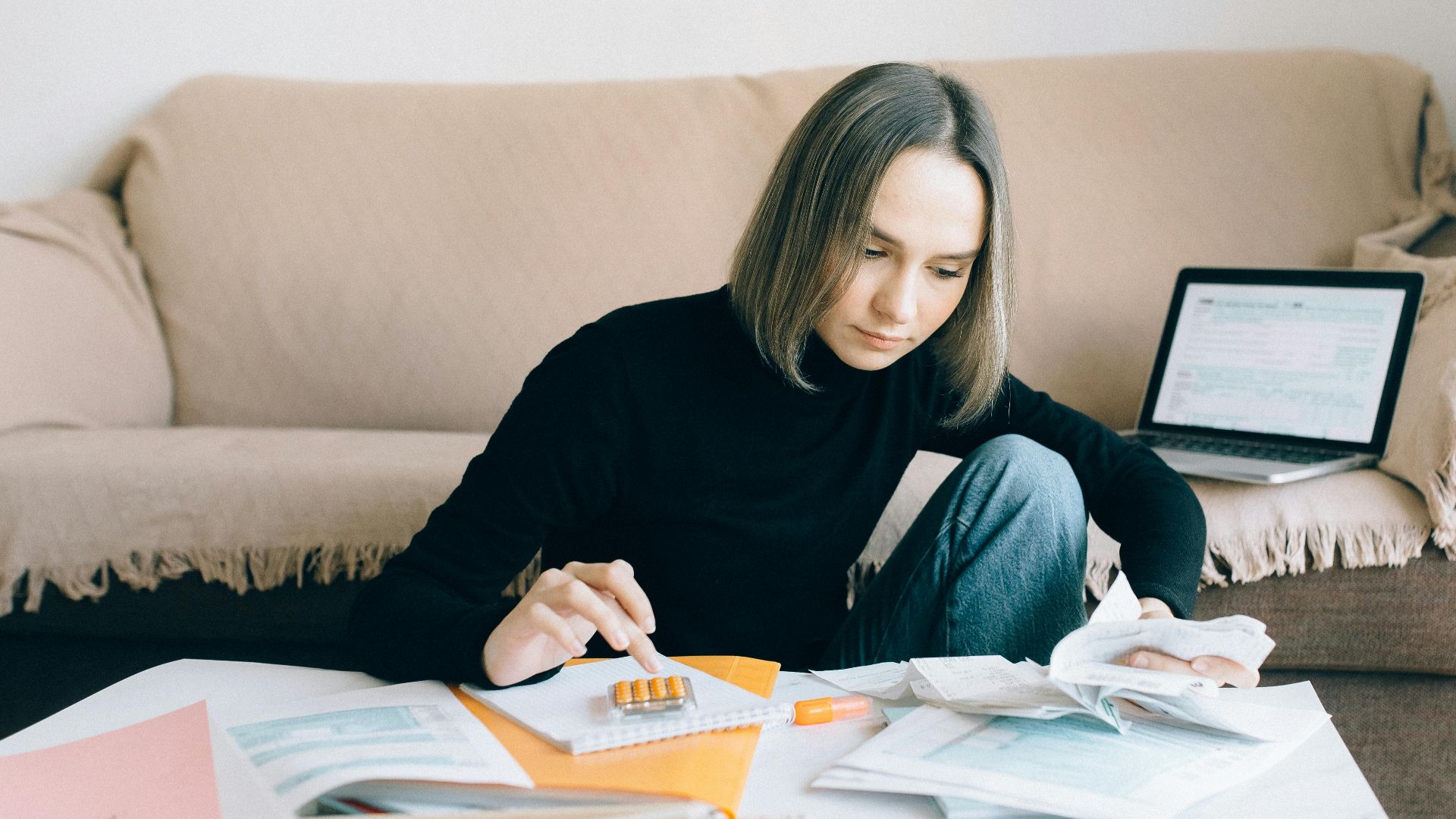 Young woman handling financial tasks with papers and laptop in cozy living room.