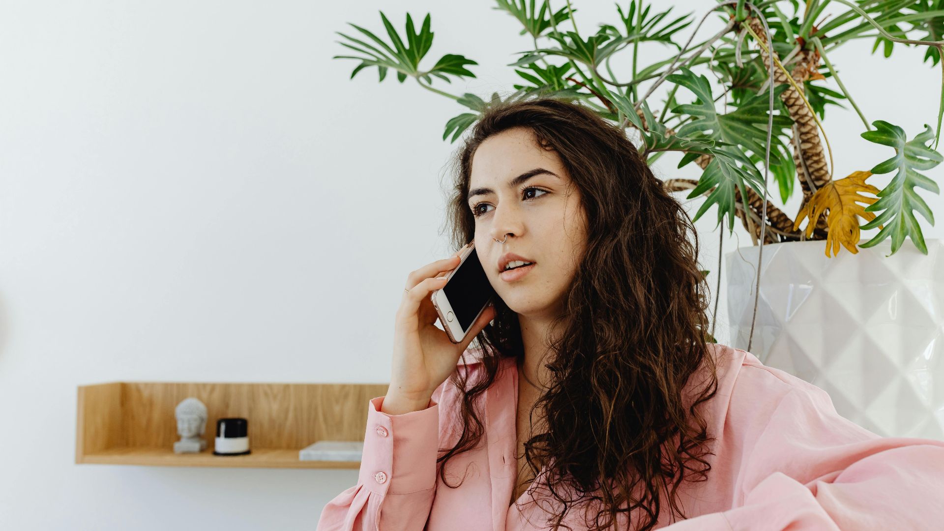 Confident young woman talking on smartphone in a modern interior with green plant background.