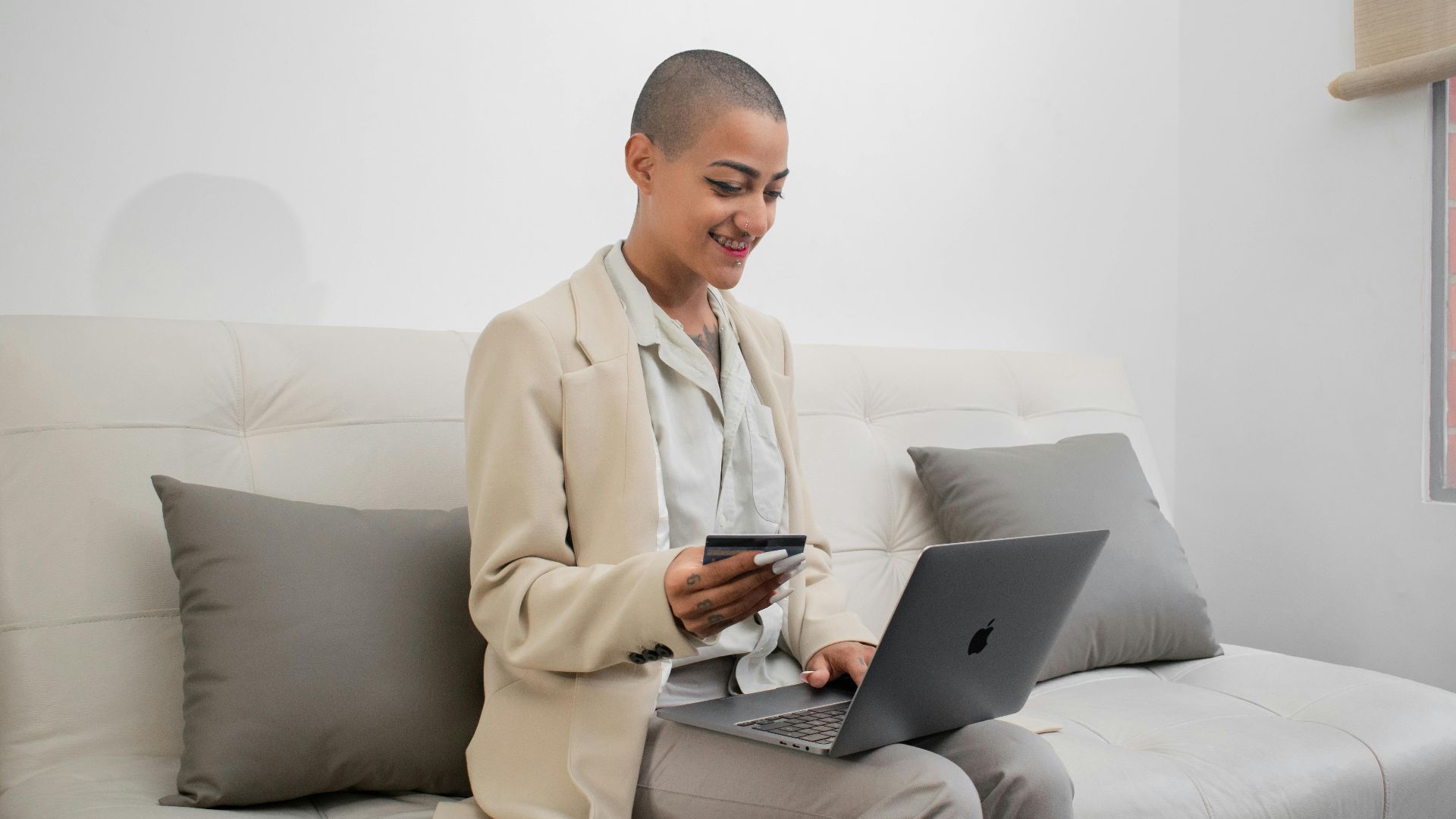 Bald woman in blazer shopping online with credit card on a couch using a laptop.