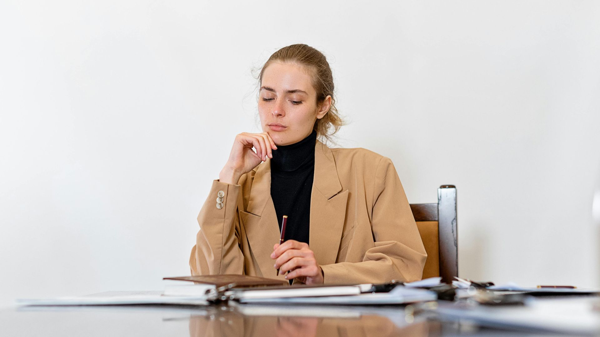 A focused young woman in a beige jacket sits at a desk with notes, deep in thought.