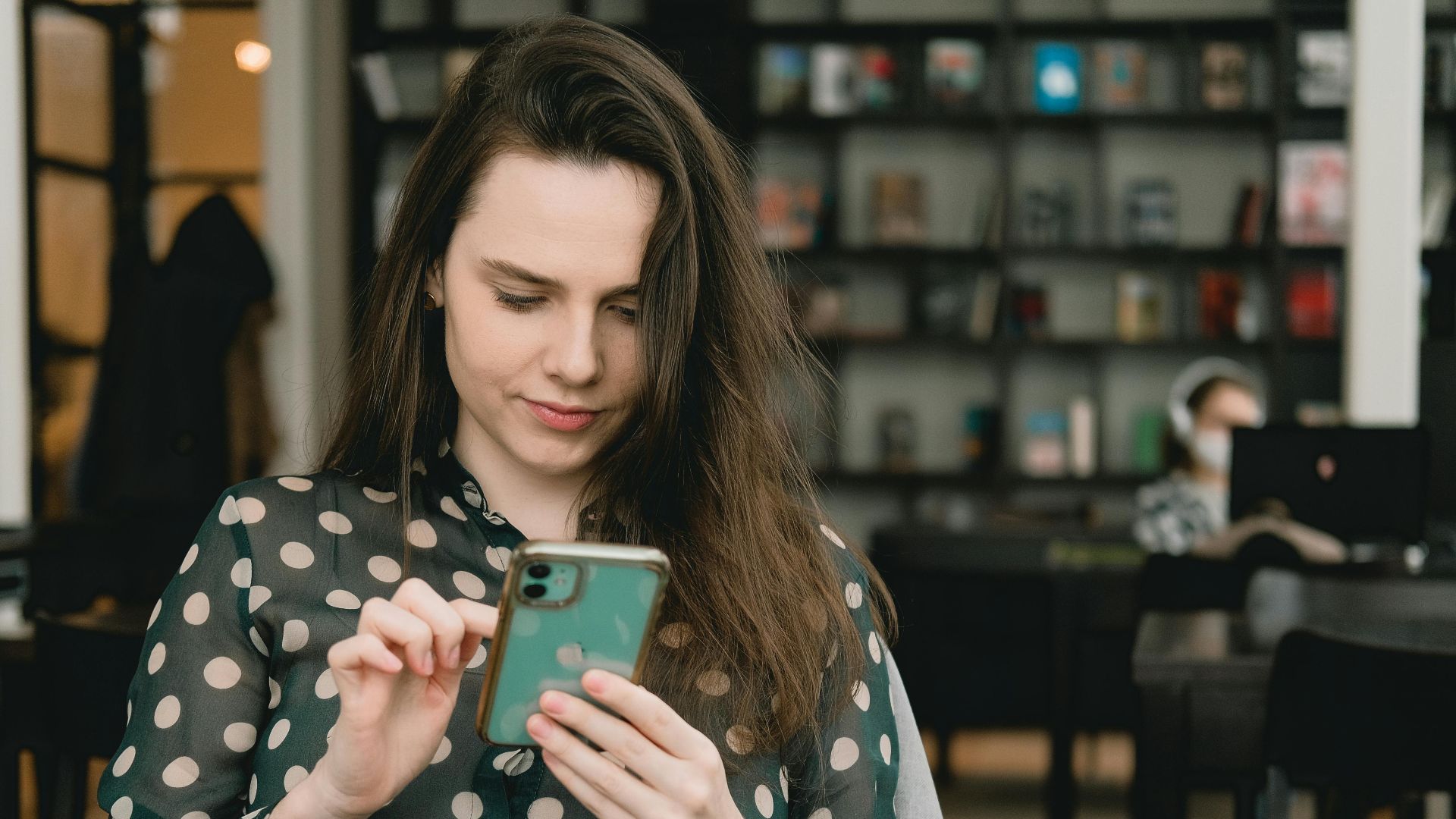 Woman in polka dot dress using smartphone indoors at a library. Cozy and modern setting.