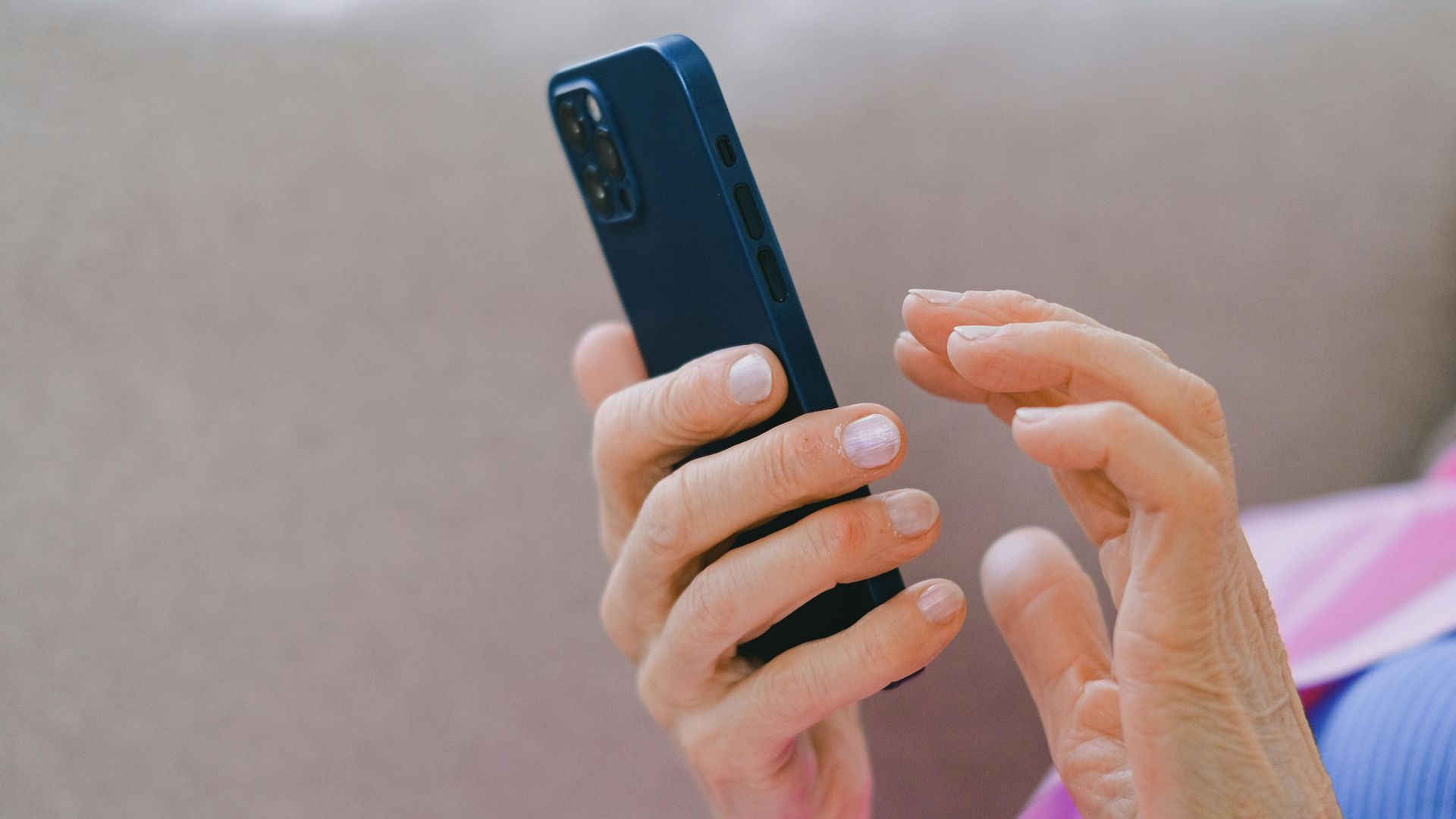 A senior adult in pink jacket using a smartphone while relaxing on a sofa indoors.