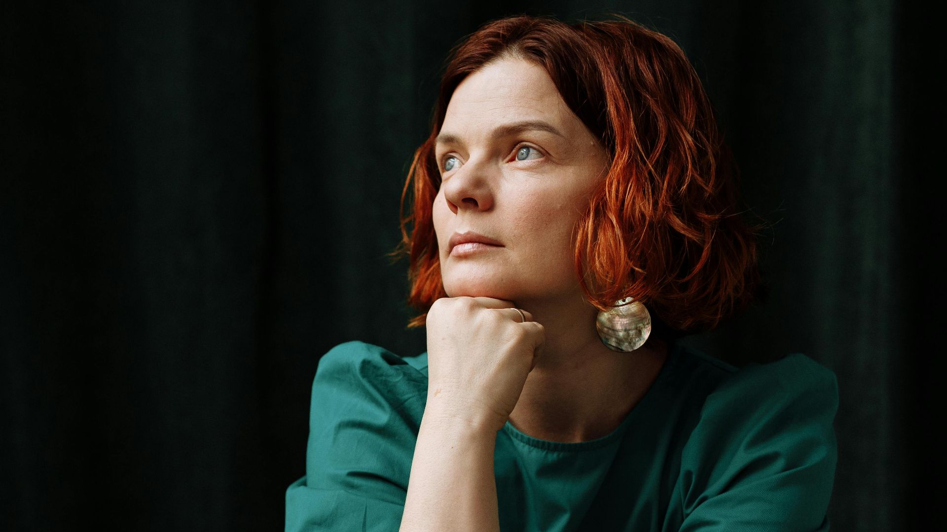 A woman in a green dress sits pensively indoors, resting her chin on her hand.