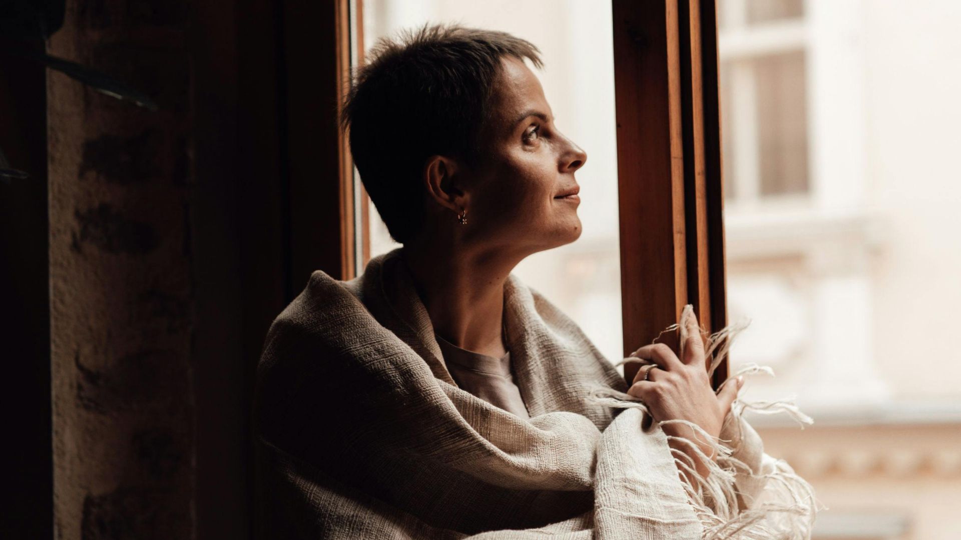 Woman wrapped in a cozy blanket sitting by a window, gazing outside.