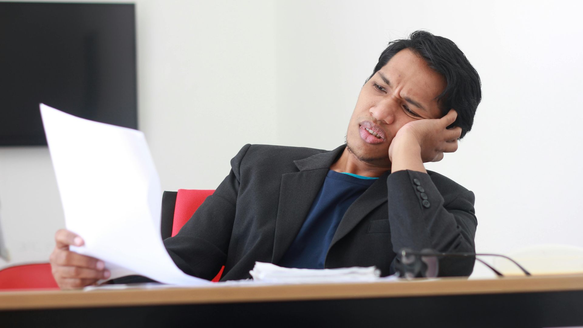 A businessman in a black suit experiences stress while reviewing paperwork in a modern office setting.
