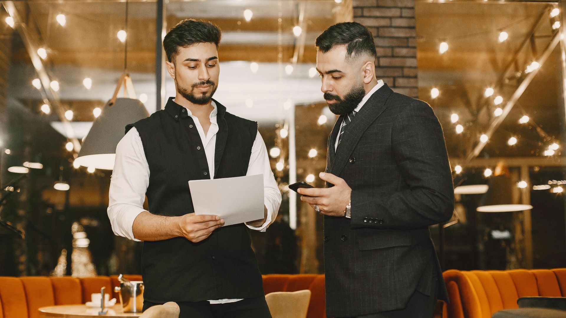 Two businessmen reviewing documents and smartphone in a restaurant setting with warm lighting.