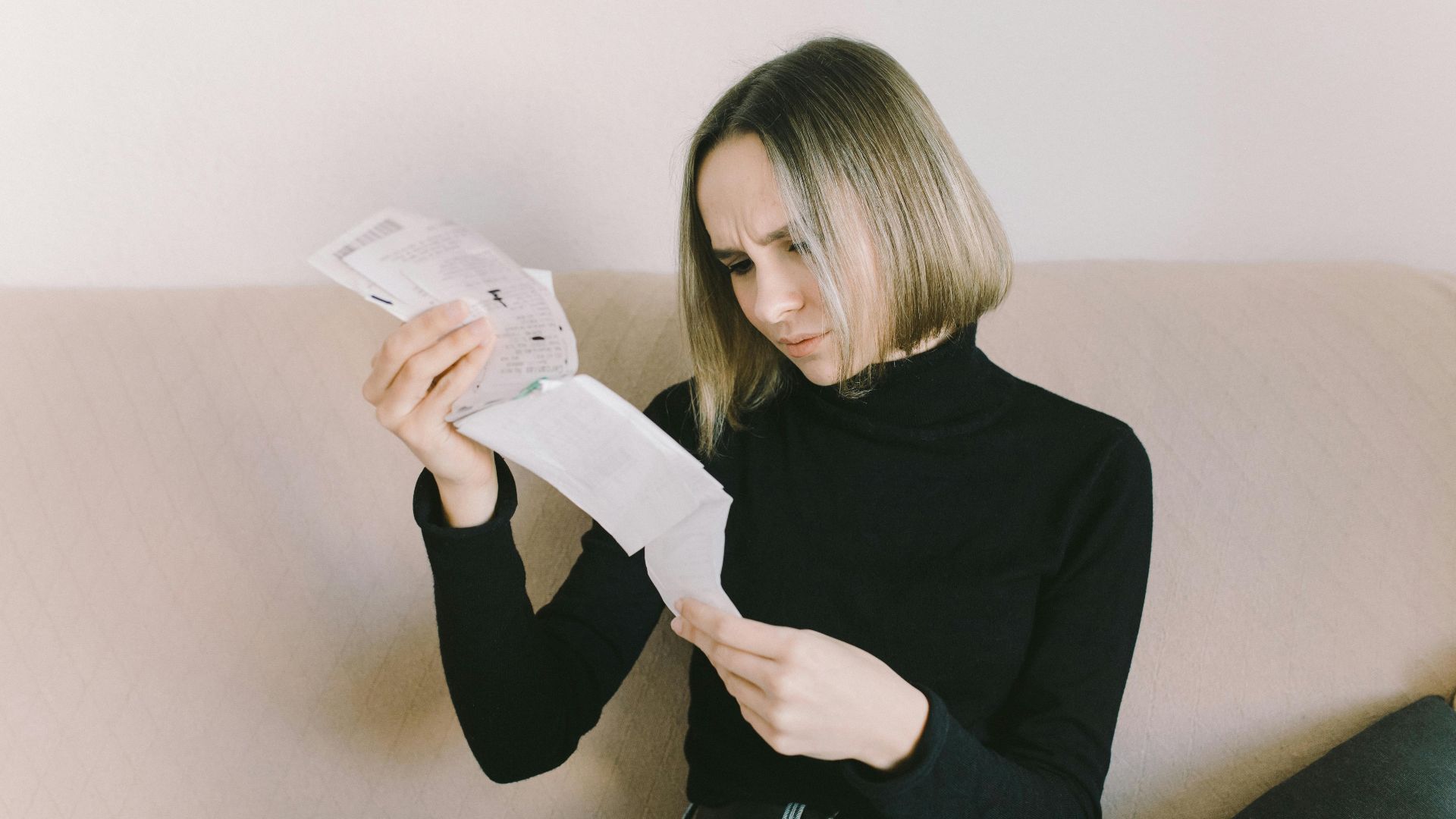 Concerned woman in black sweater examining bills on beige sofa indoors.