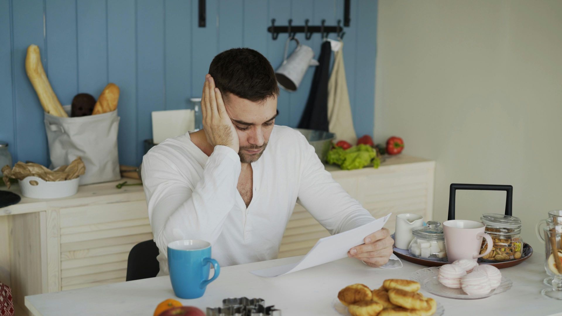 Man in white shirt reading a document at kitchen table with breakfast items.