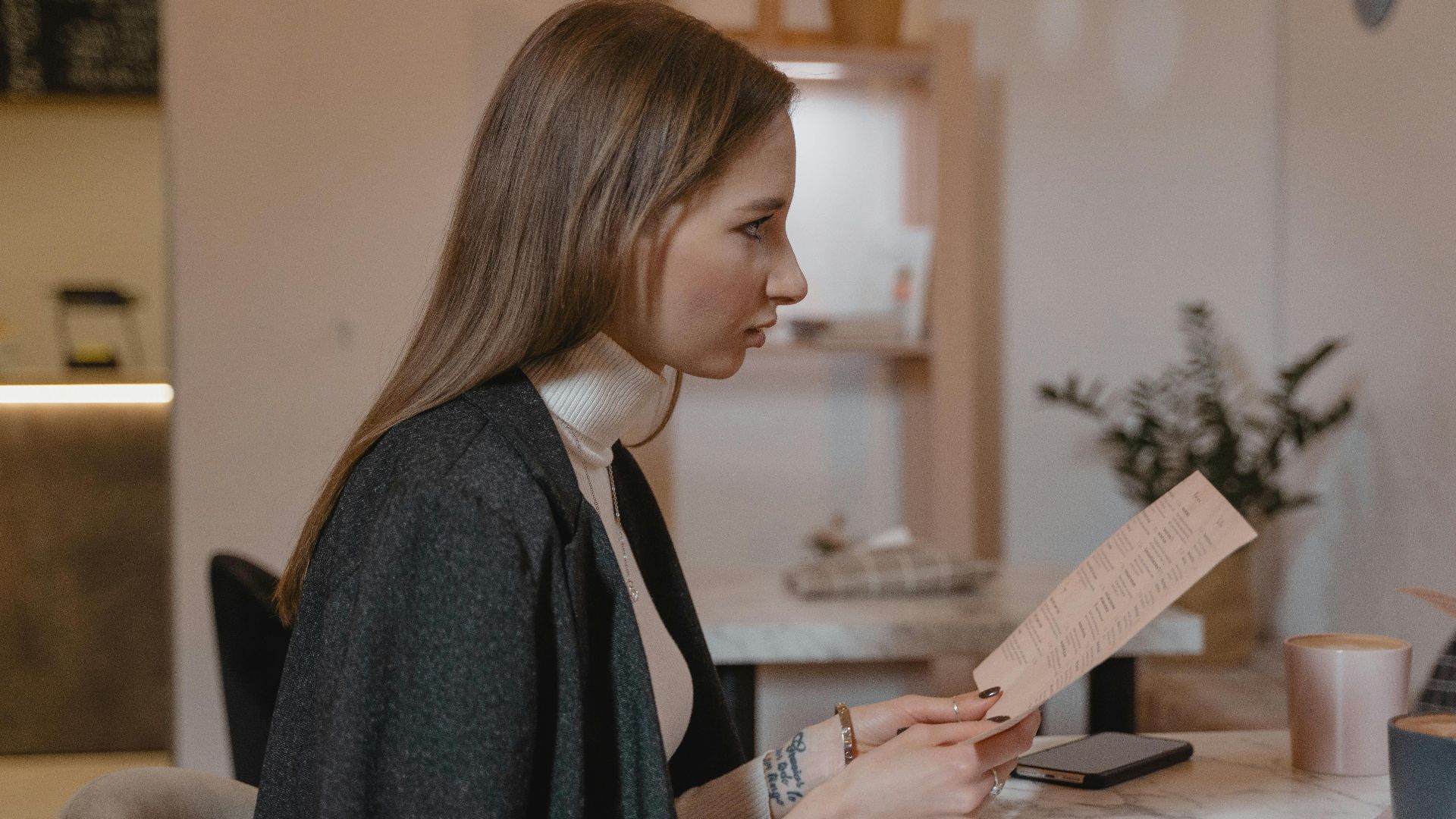 Side view of woman holding menu in a cozy café setting, wearing a turtleneck and jacket.