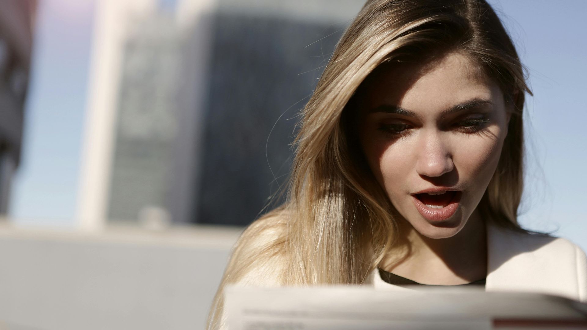 A young woman with blonde hair expresses surprise while reading a newspaper outdoors in daylight.