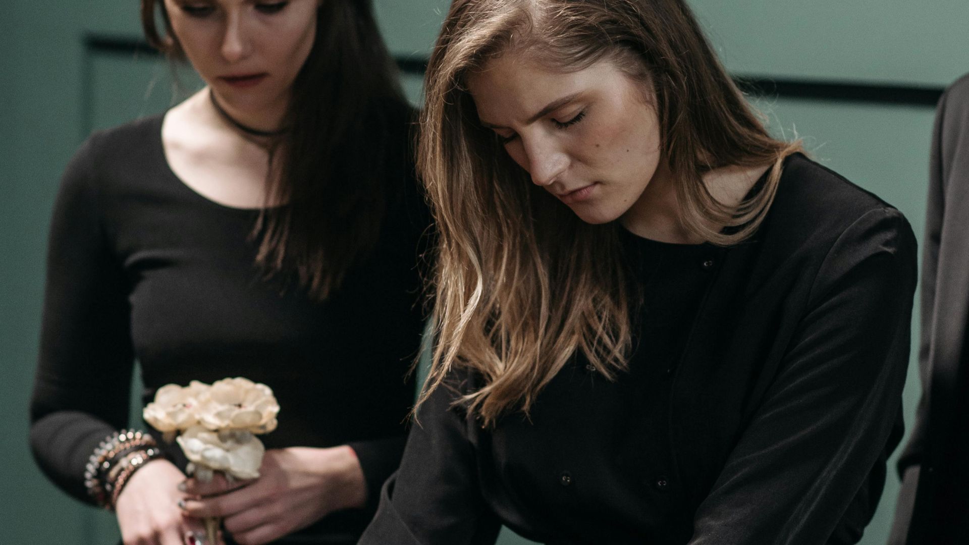 Two women expressing grief by a coffin, holding flowers in a solemn funeral setting.