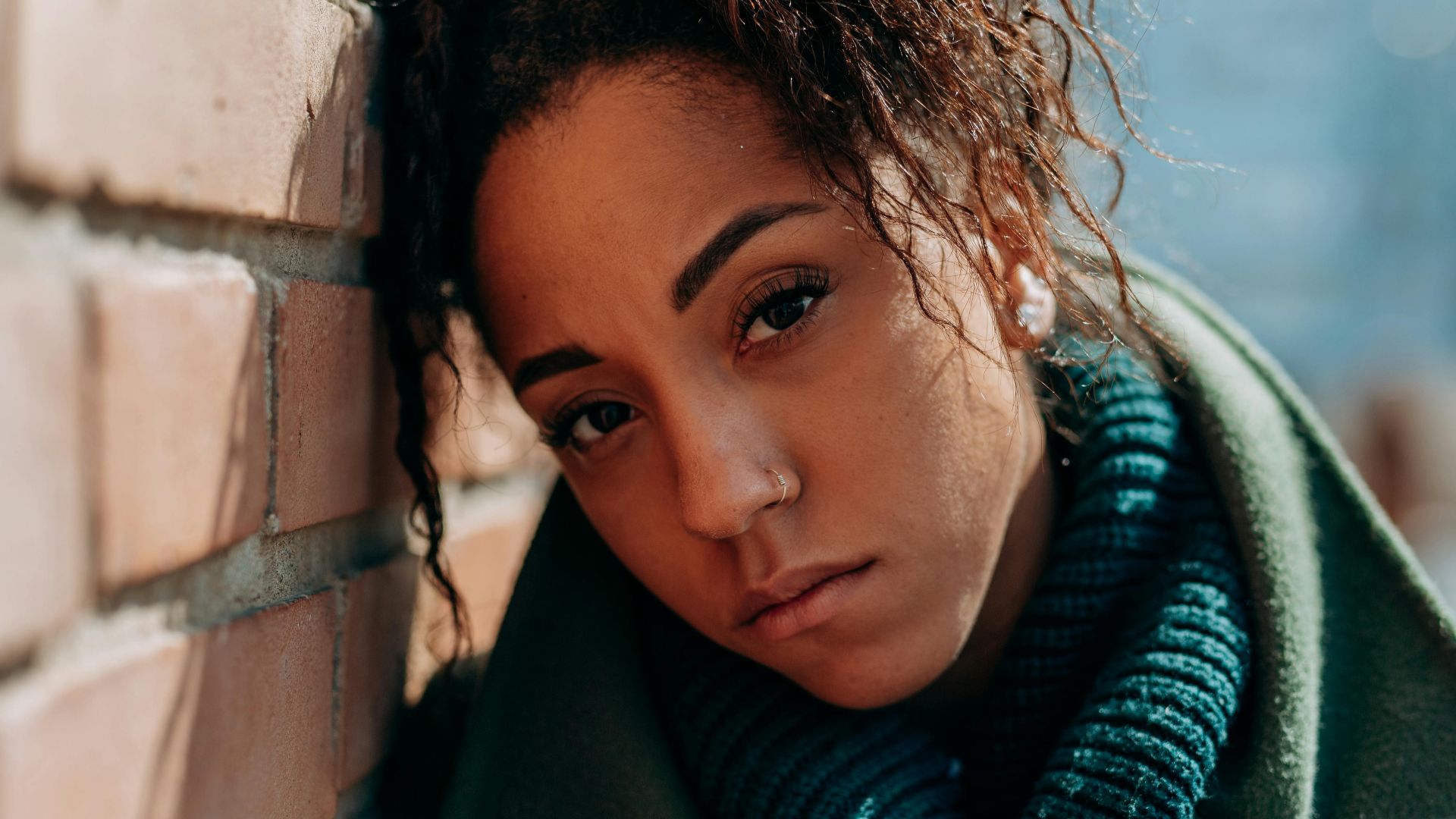 A thoughtful woman in a green coat leans against a brick wall on a sunny day.