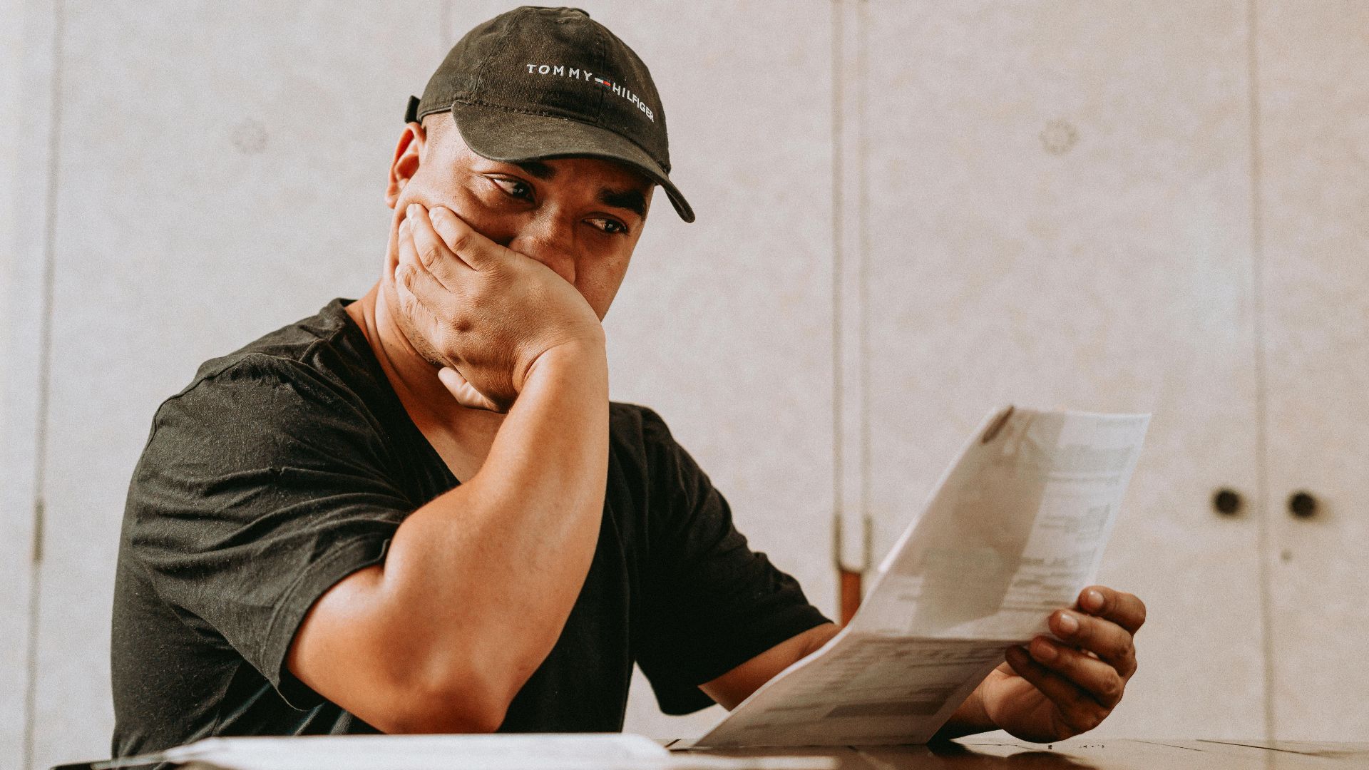 A man sits indoors, deeply focused on reviewing paperwork, expressing concern and contemplation.